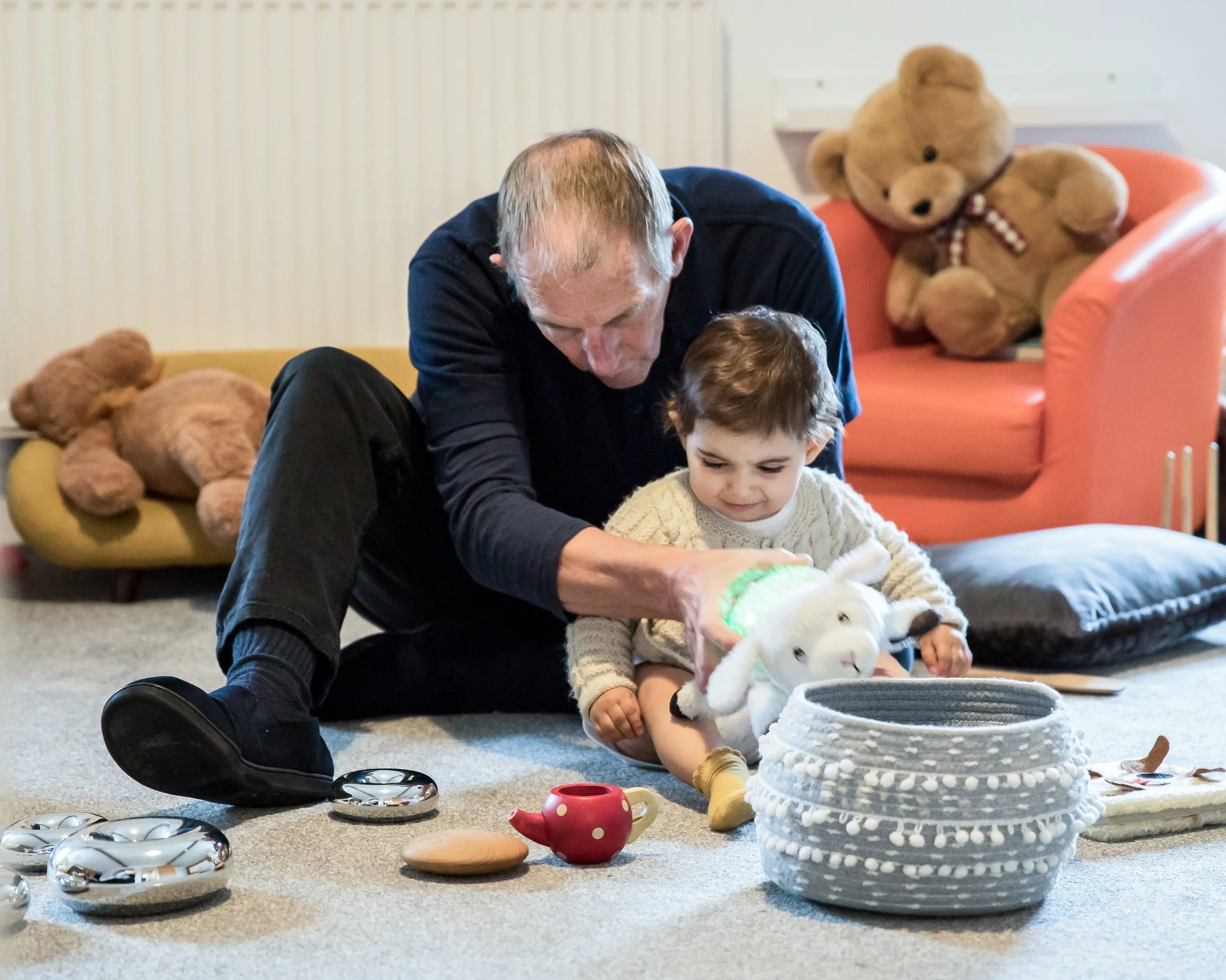 An adult and a young child sitting on the carpeted floor playing with a stuffed bunny toy, surrounded by various toys and a large white woven basket, with teddy bears on the couches in the background.
