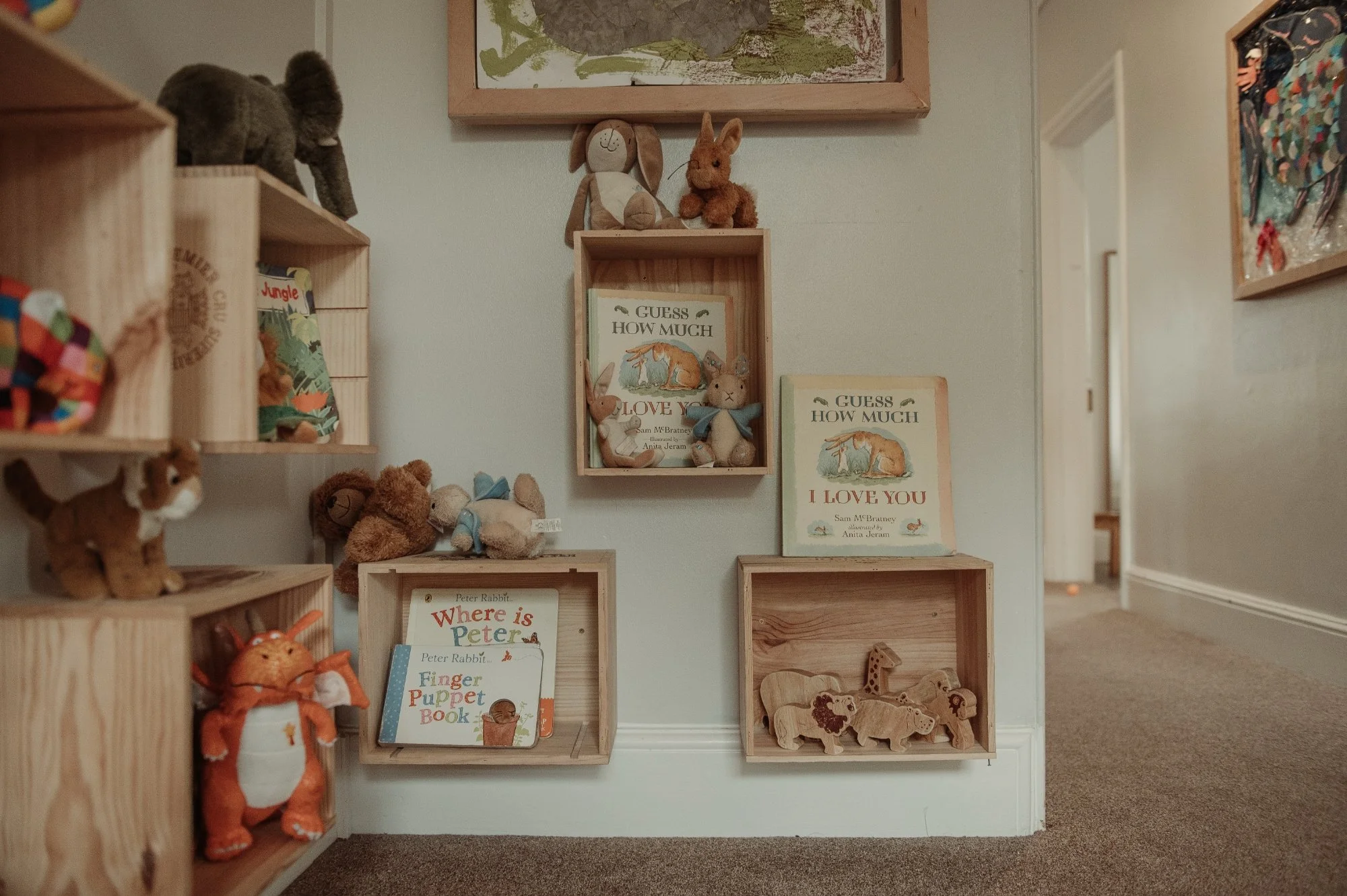 A corner of a children's room with wooden shelves and boxes holding stuffed animals, children's books, and wooden animal toys. Decor includes plush rabbits, a dog, lion, elephant, and bird dolls, with framed artwork on the wall.