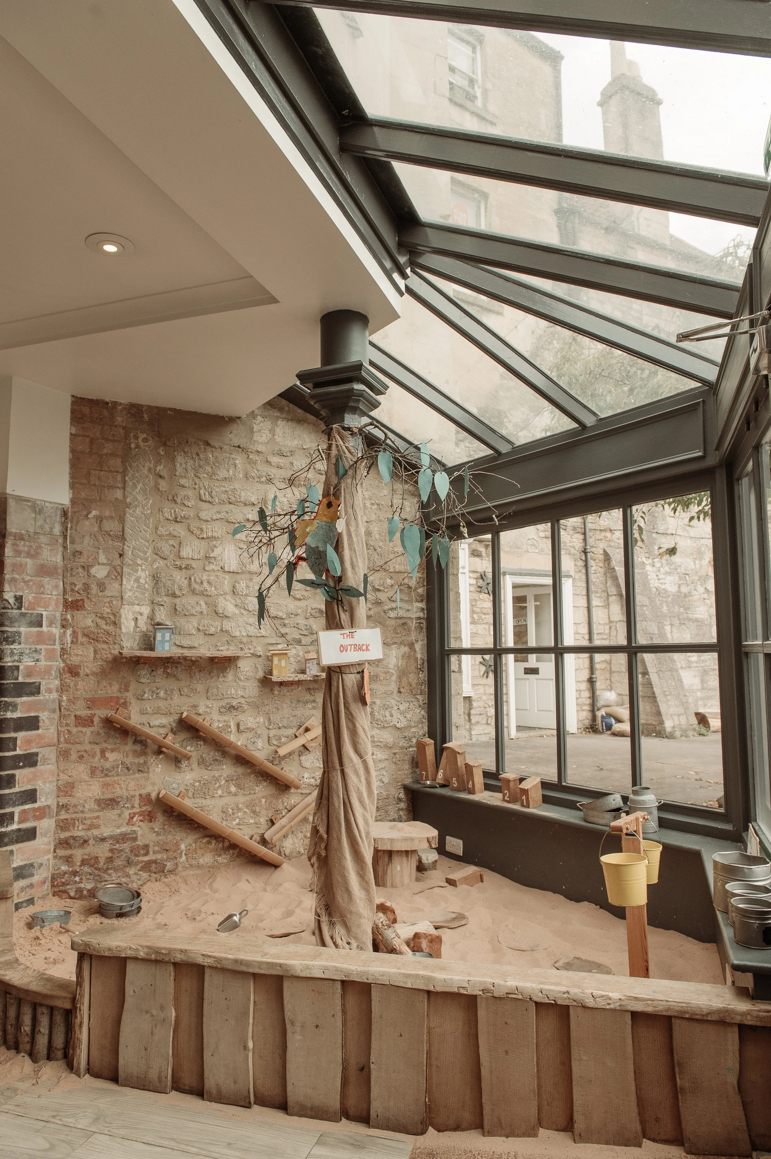 Indoor space resembling a play area with sand, wooden structures, and a tree branch decoration under a large glass roof.