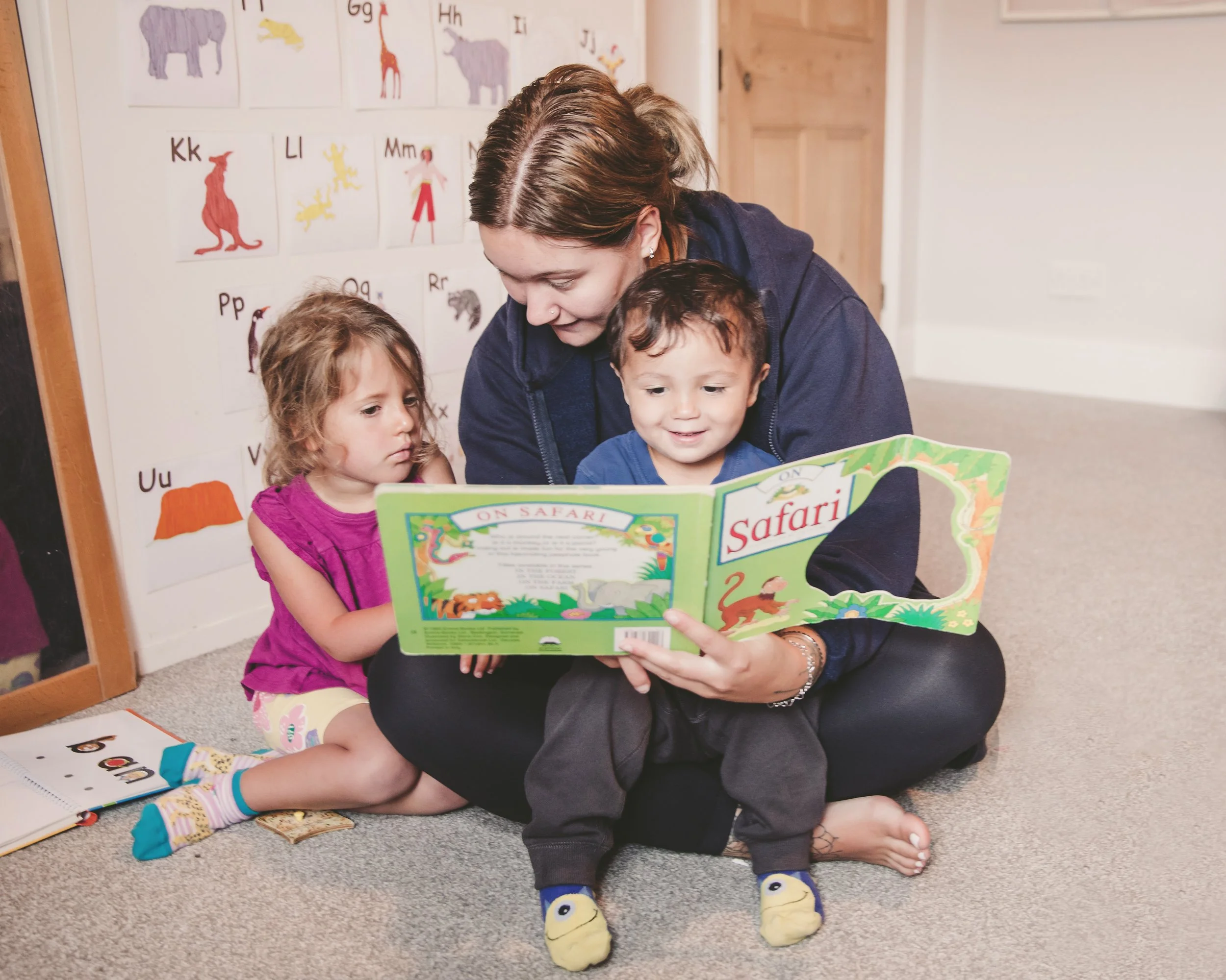 A woman reading a children's book titled "On Safari" to two young children, a boy and a girl, in a room with educational animal and alphabet posters on the wall.