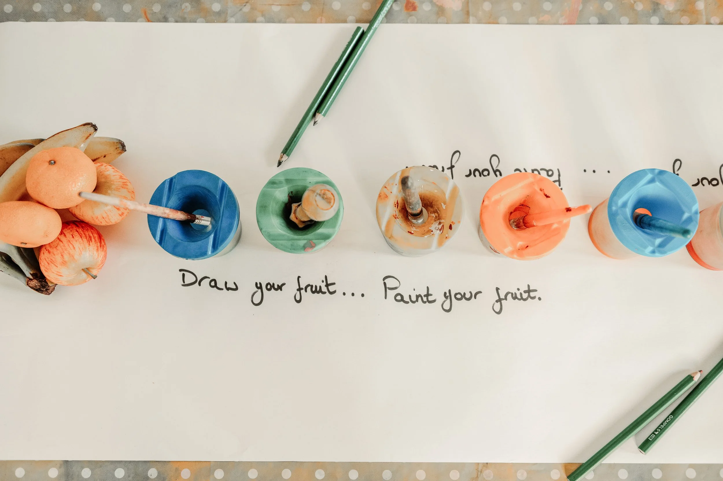 Craft activity with artificial fruit, paint, and brushes on a white paper with hand-written instructions reading 'Draw your fruit... Paint your fruit.'