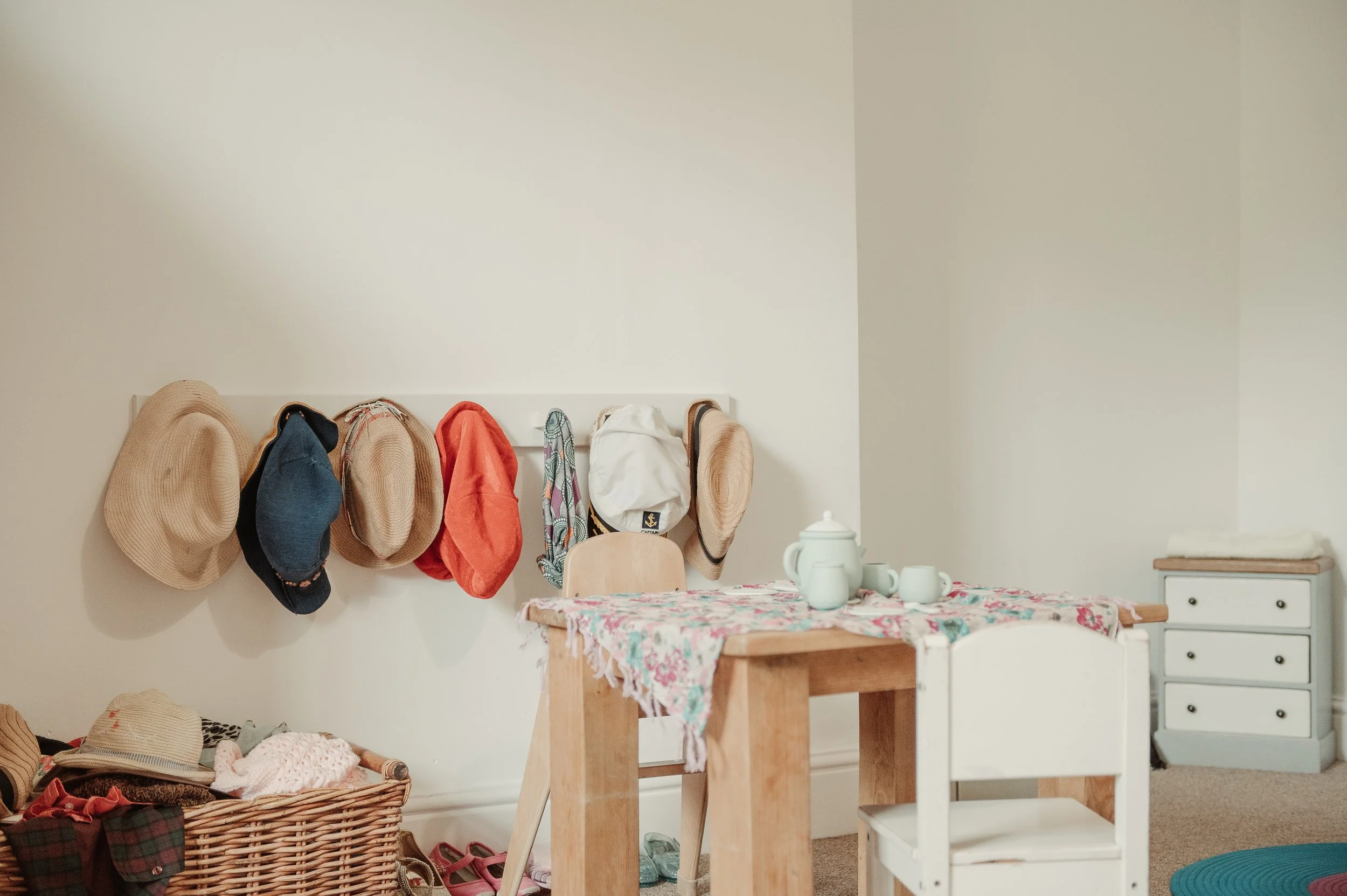A white wall with hats hanging on a rack, a basket with clothes and hats on the floor, a wooden table with a floral tablecloth and a tea set, and a small dresser with a stack of cloth on top.