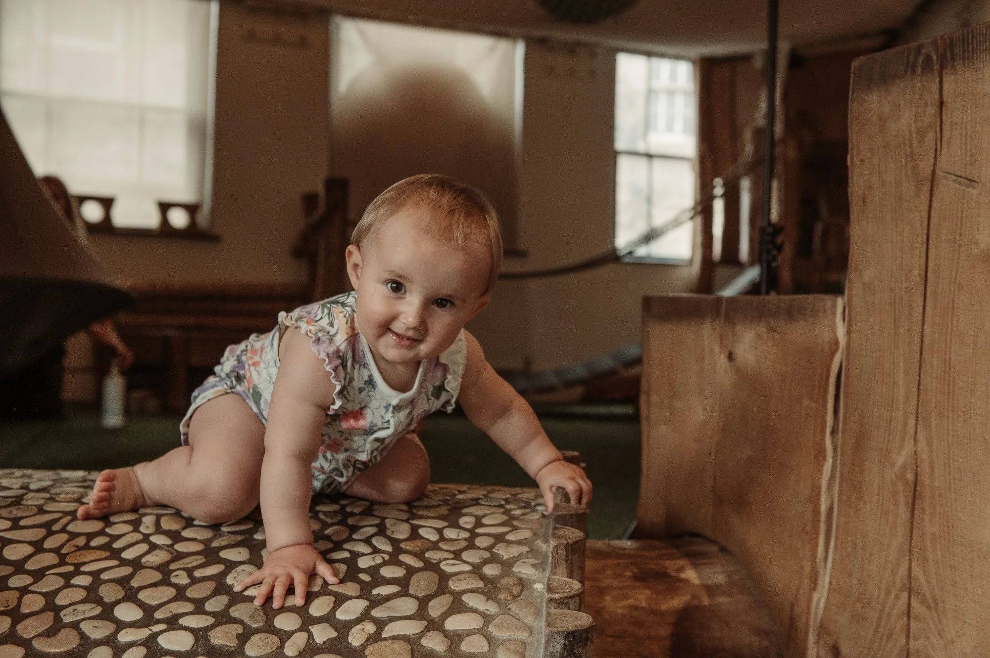 Baby crawling on a pebble-patterned surface indoors with wooden furniture and windows in the background.