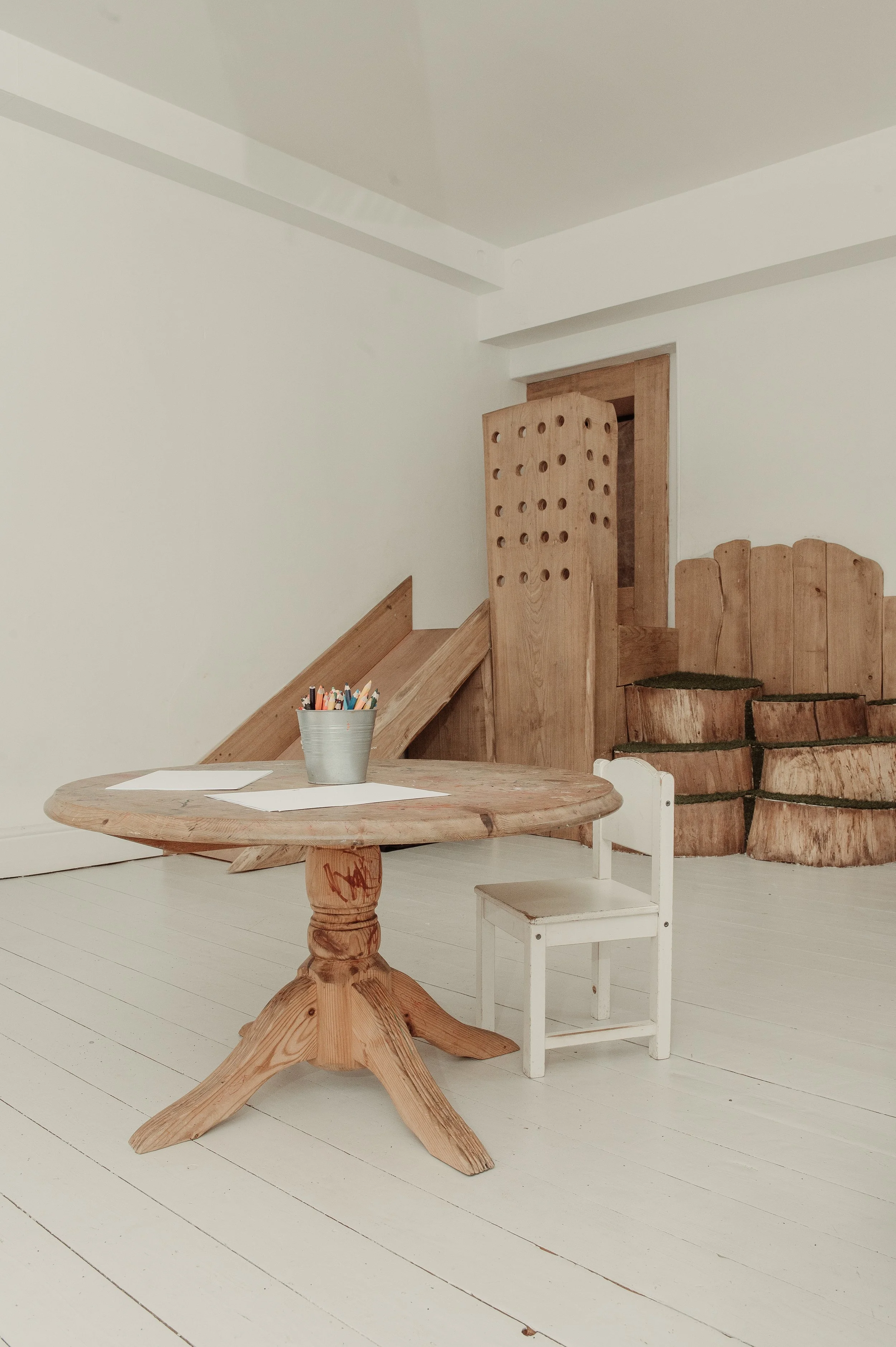 A wooden children's table with a white chair, a bucket of colored markers, and old wooden barrel seats in the background.