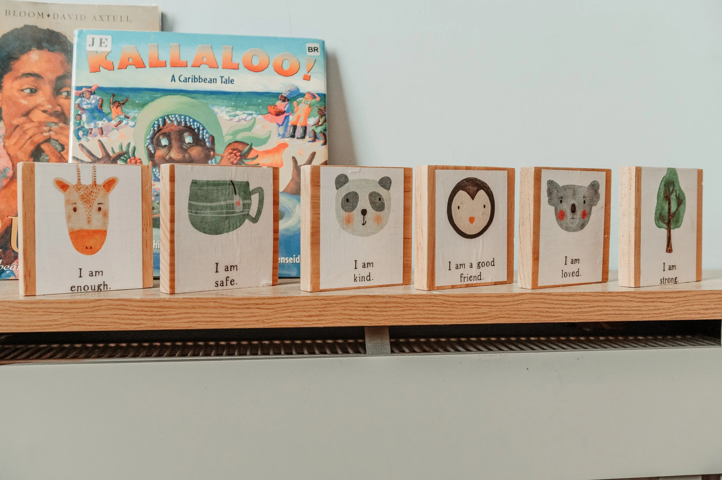 Six wooden blocks with illustrated animals and positive affirmations on a shelf, with books in the background.