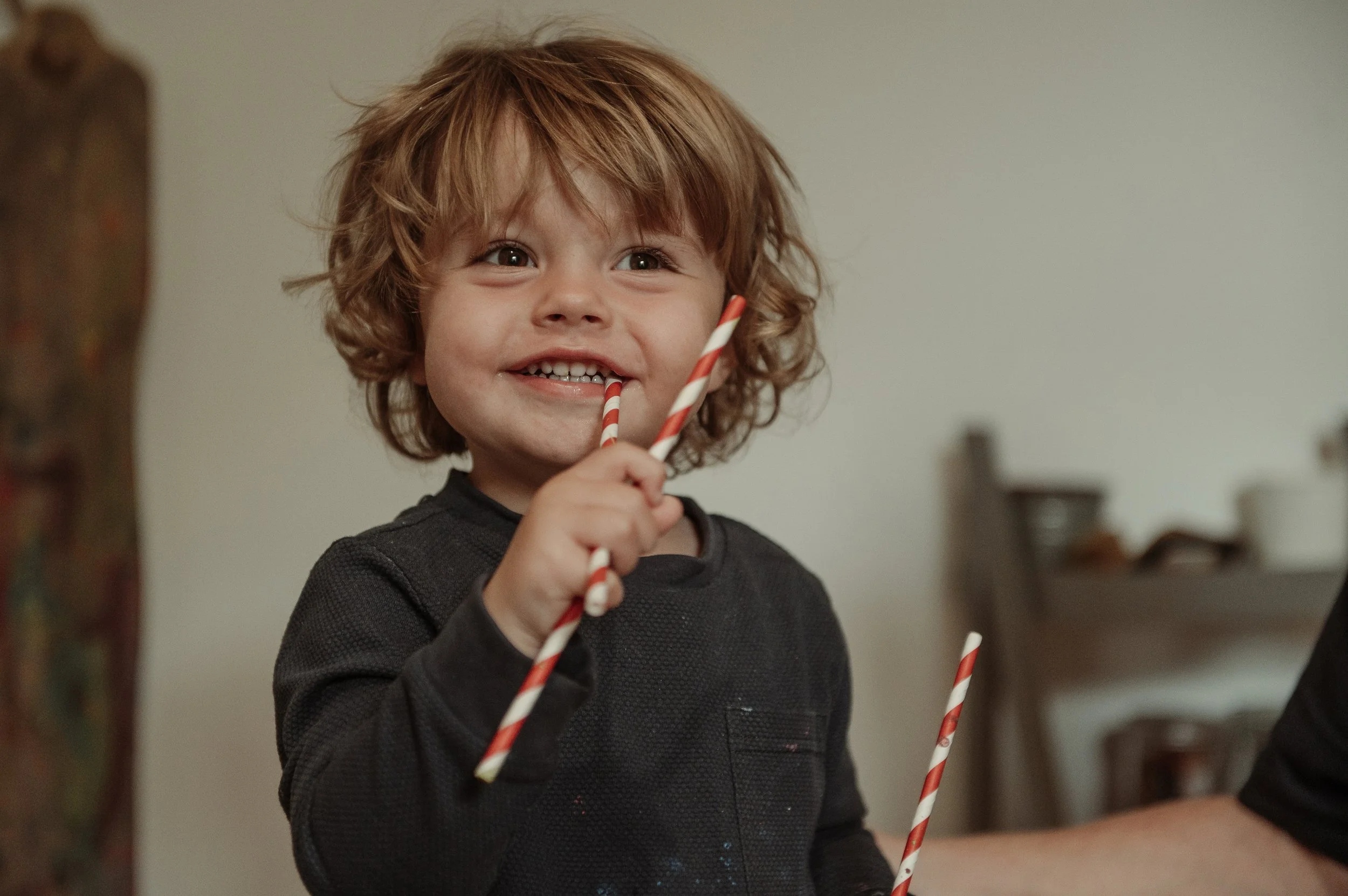 A young boy with curly red hair smiling and holding two red and white striped straws.