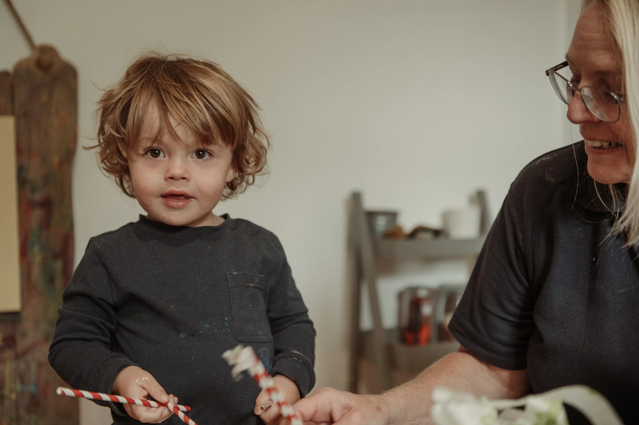 A young boy with curly red hair and a woman with glasses smiling, holding red and white striped candle sticks in a room with a wooden shelf in the background.