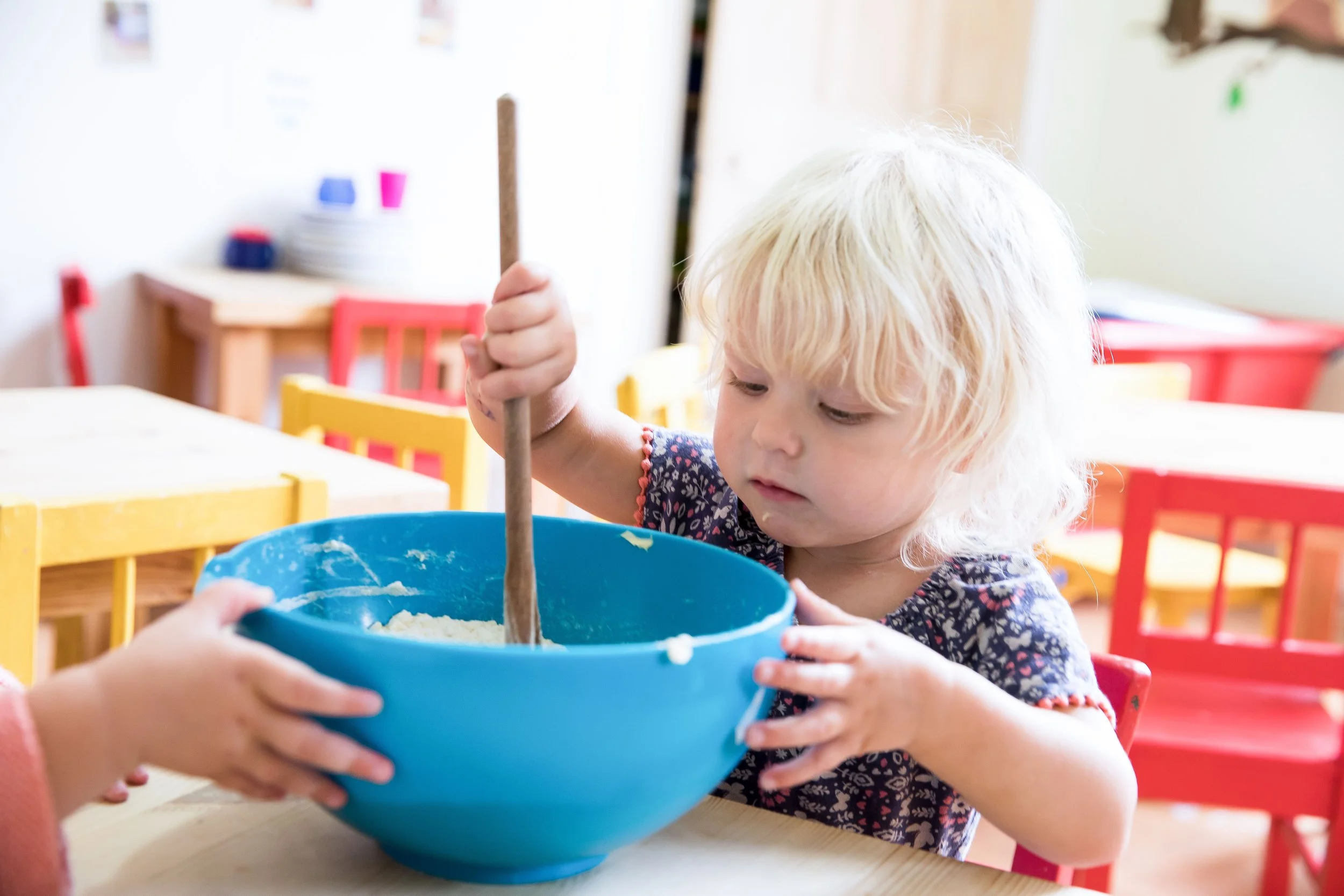 A young child with blonde hair stirring a mixture in a large blue bowl, with another child's hand helping to hold the bowl, seated at a table in a colorful classroom or play area.