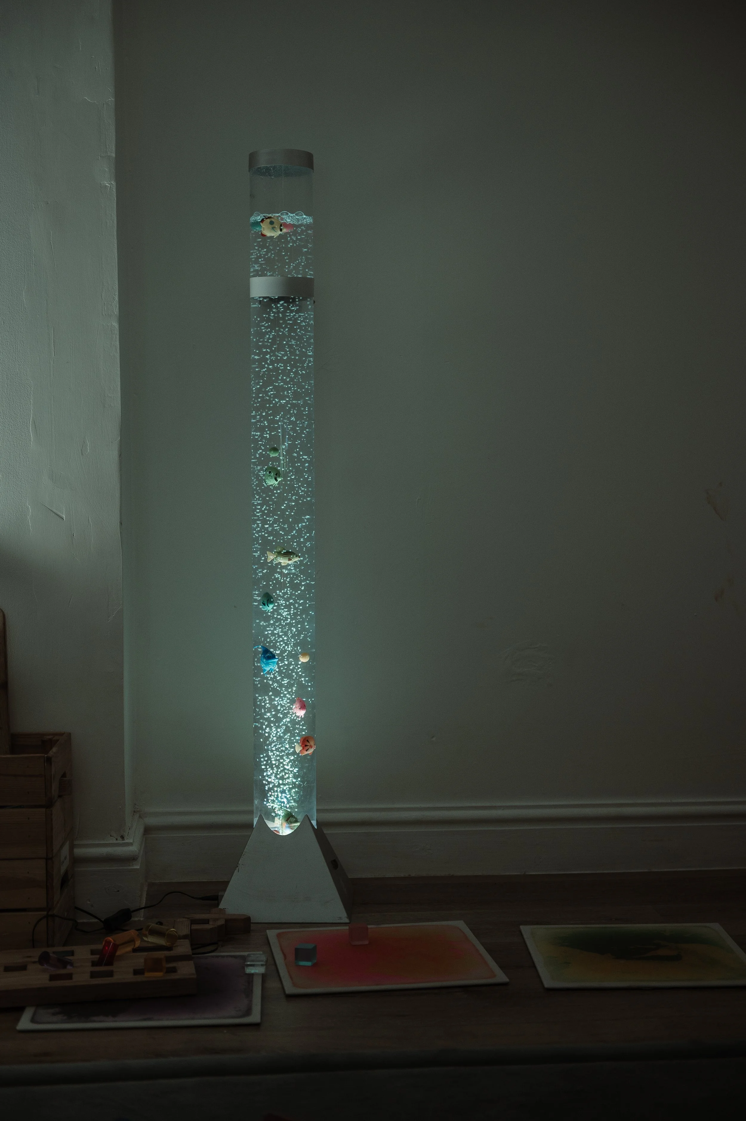 A tall, illuminated, transparent bubble tube with colorful fish-shaped ornaments inside, placed on a wooden surface in a dimly lit room.