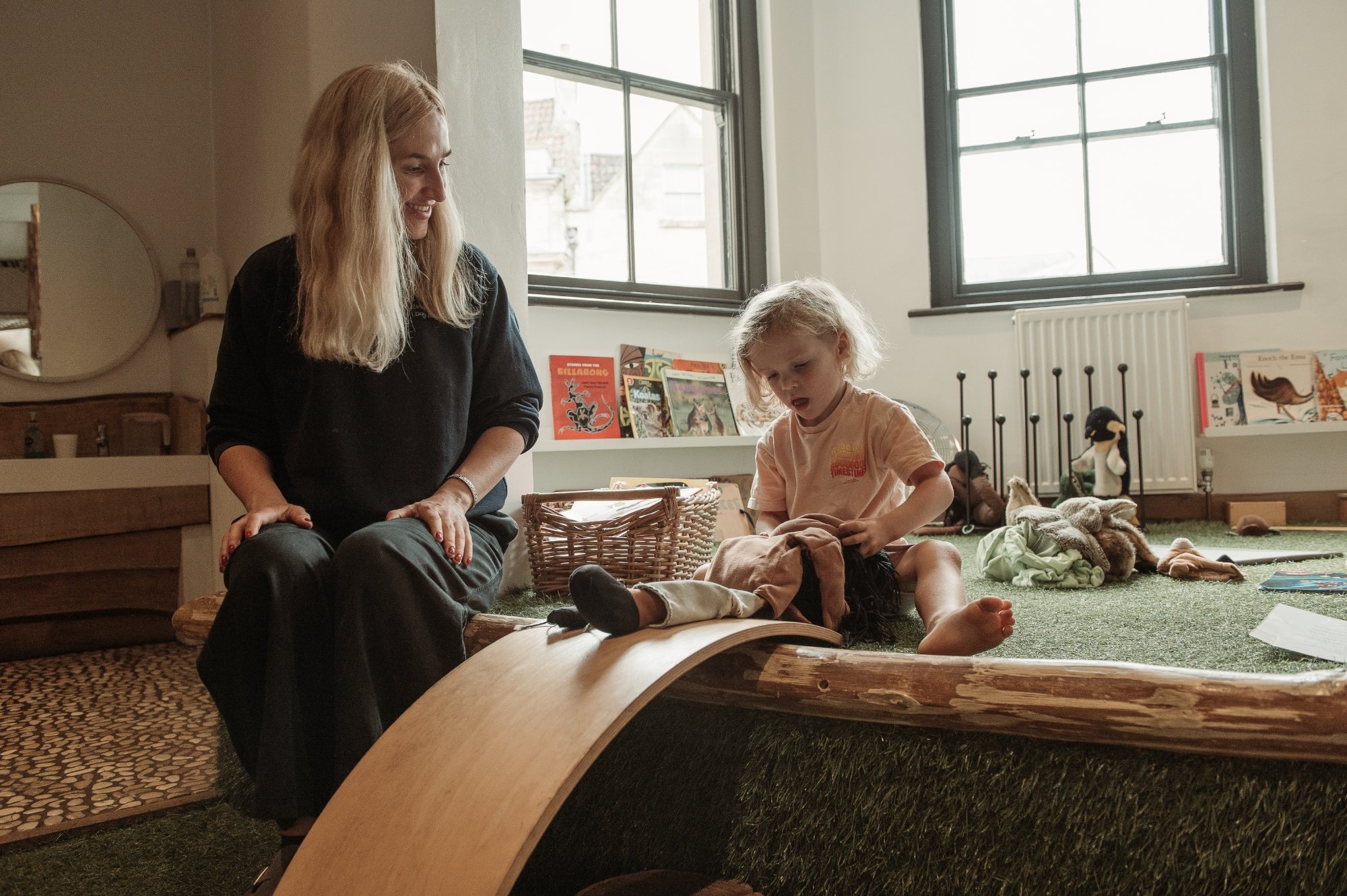 A woman and a young girl playing indoors on a curved wooden ramp. The woman is watching and smiling, while the girl is focused on her clothes. There are books and plush toys in the background.