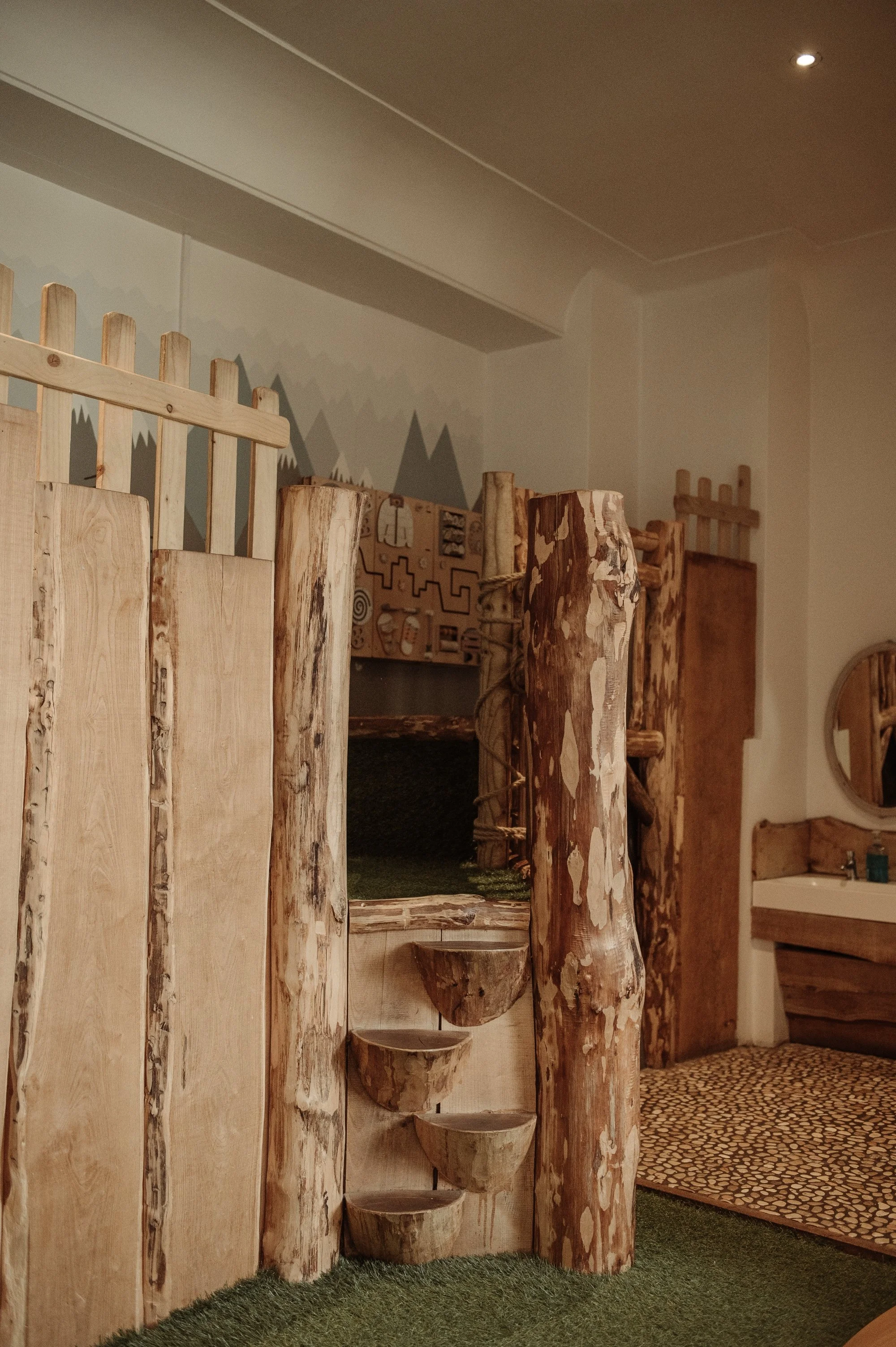 Indoor play area with wooden tree trunk structures, climbing steps, and a small wall with puzzle pieces; natural wood and greenery decor.