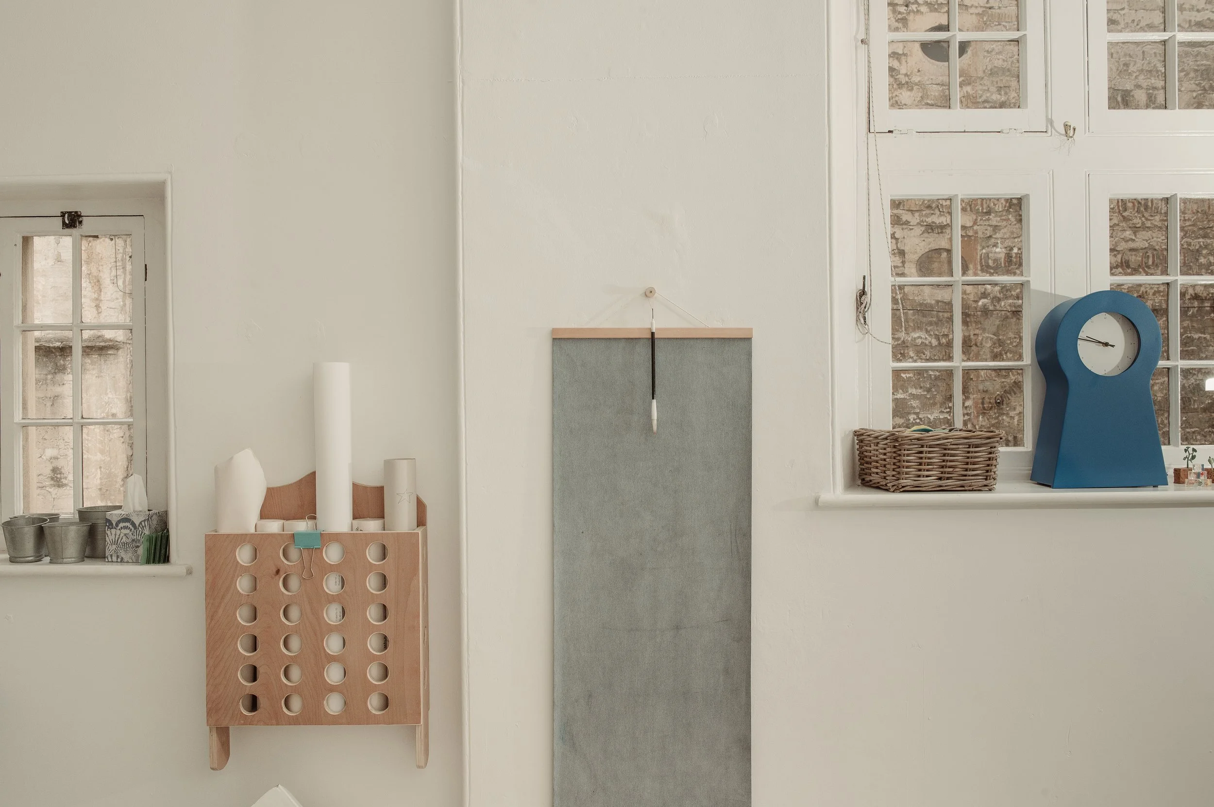 Minimalist interior with white walls, a window, a grey hanging board, a blue clock on the windowsill, and small potted plants.