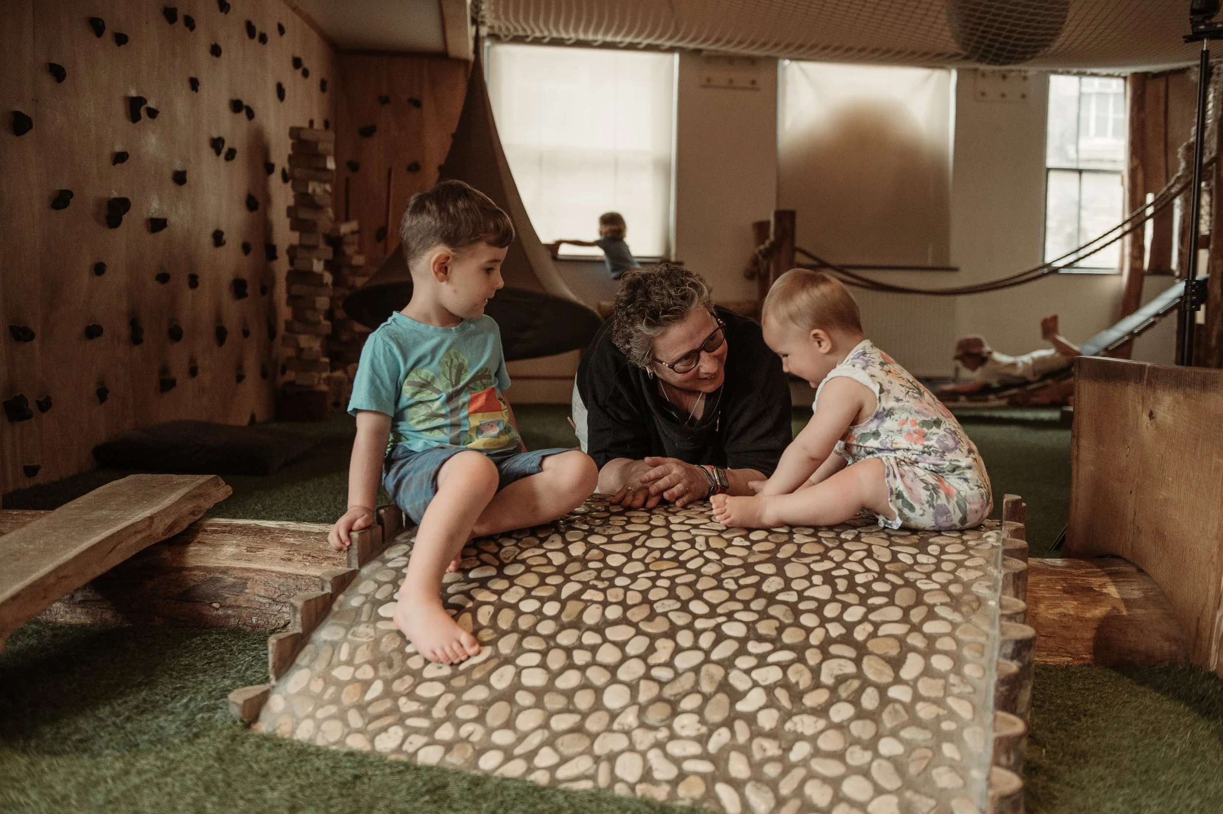 A woman with glasses playing and talking with two young children, a boy and a girl, on a pebble mosaic table inside a cozy indoor play area.