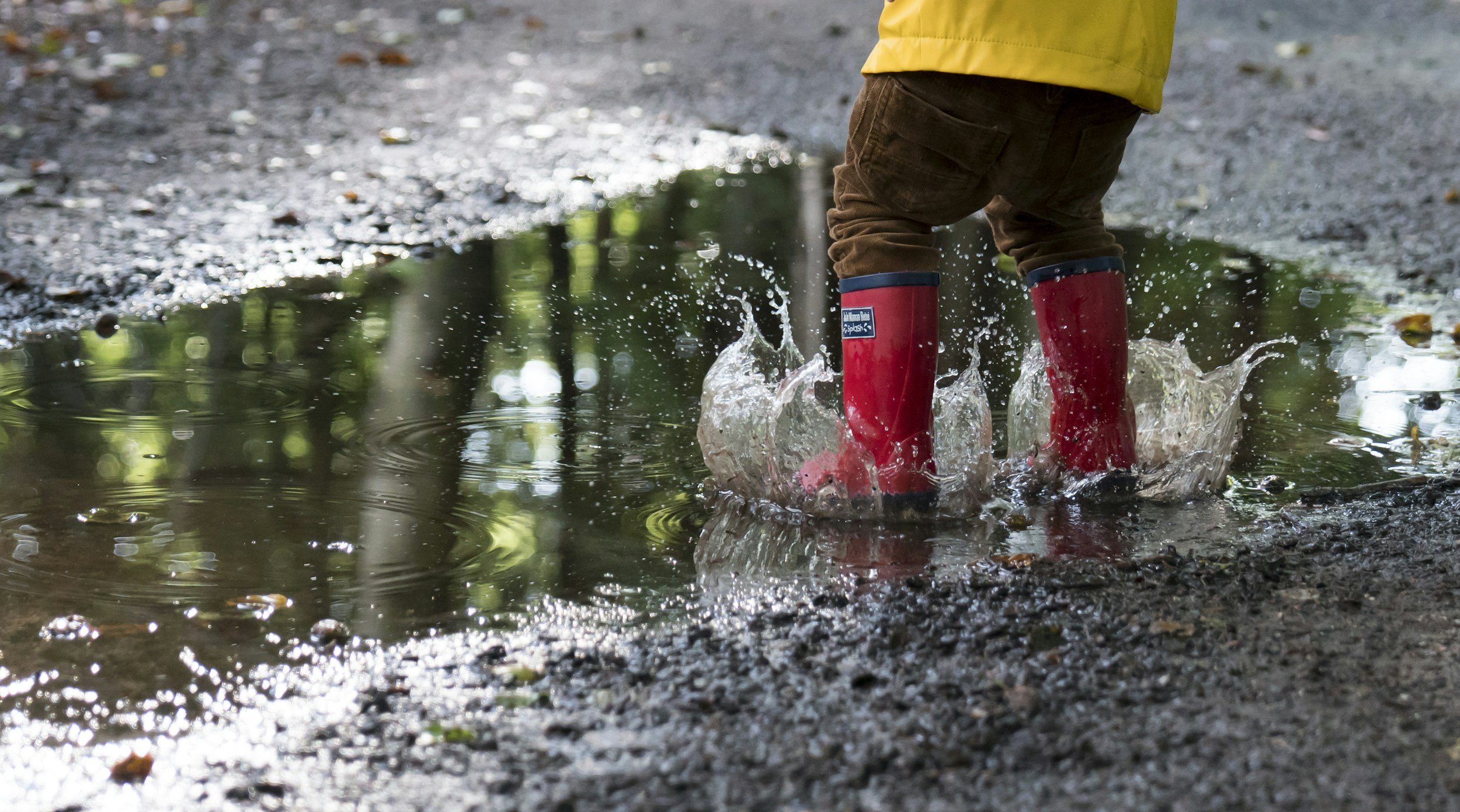 Child wearing red rain boots and brown pants splashing through a puddle on a gravel path, with a yellow raincoat visible from behind.