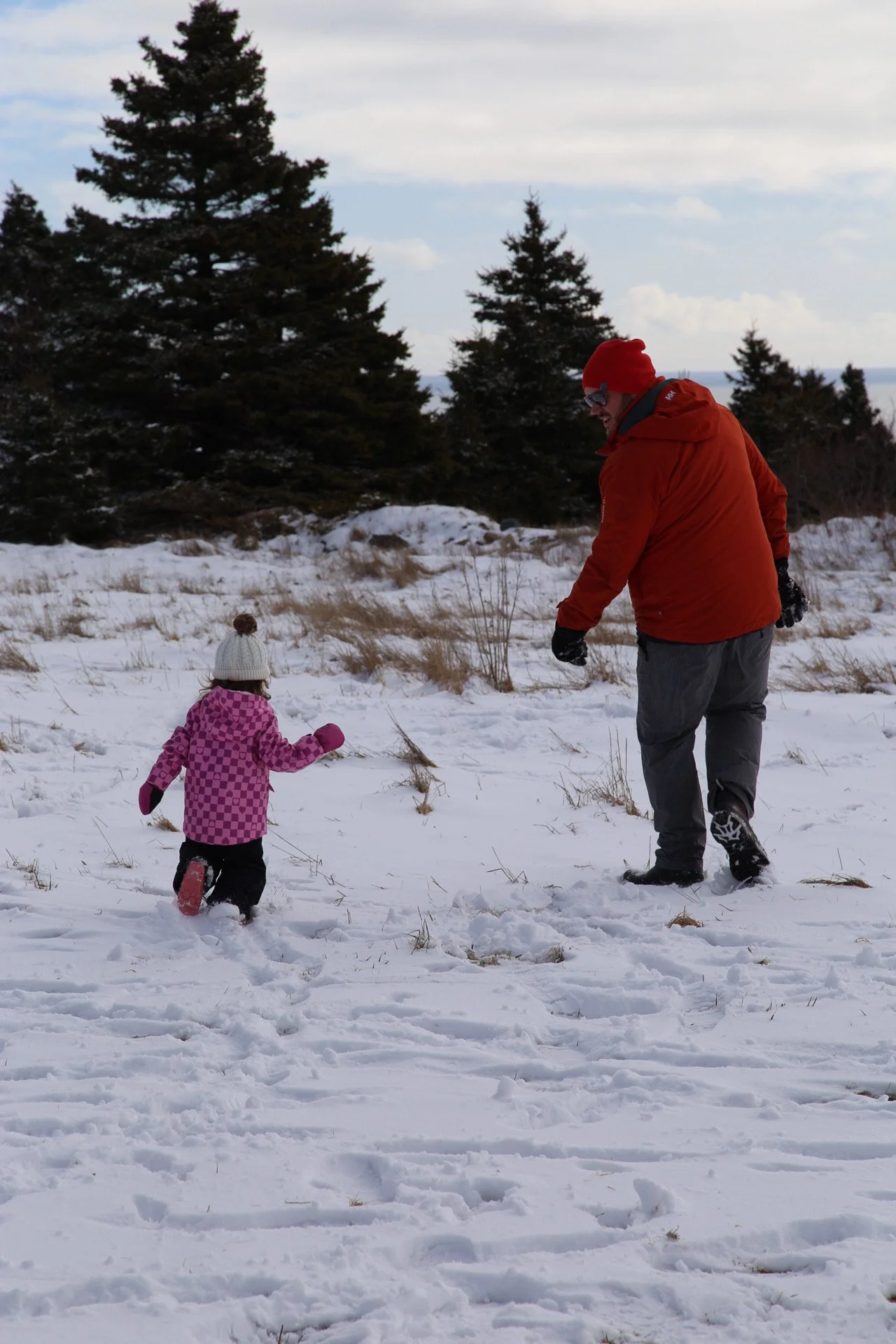 A man and a young girl playing in the snow outdoors with evergreen trees in the background. The man is dressed in a red jacket, gray pants, red beanie, and glasses, while the girl wears a pink checkered jacket, black pants, white knit hat with a pom-pom, and pink gloves.