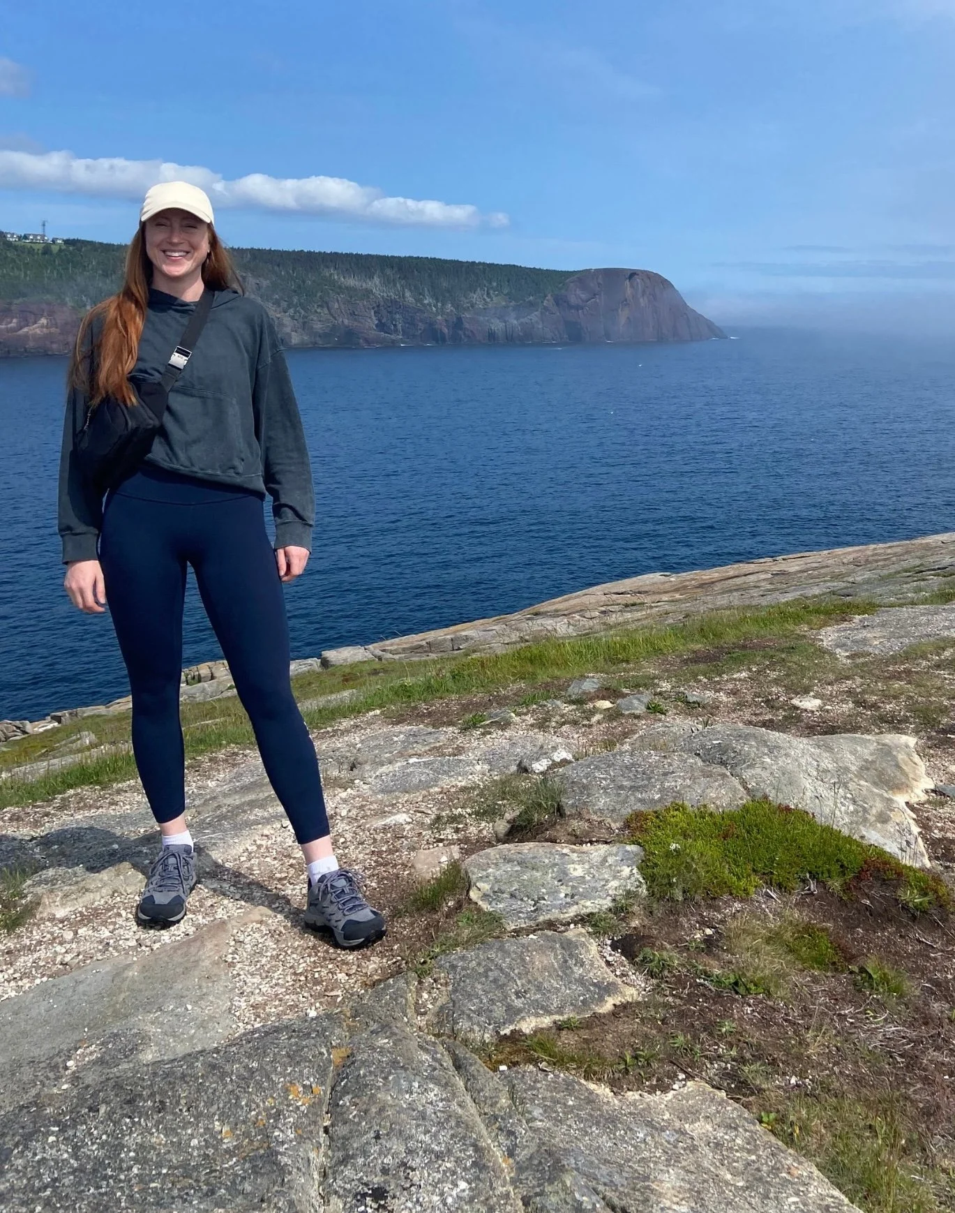 A young woman with long red hair, smiling, standing on rocky terrain near a body of water with cliffs in the background on a clear, partly cloudy day.