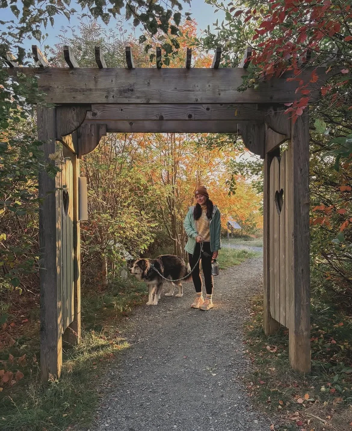 A woman with a dog walking through a wooden gate on a trail surrounded by autumn trees.