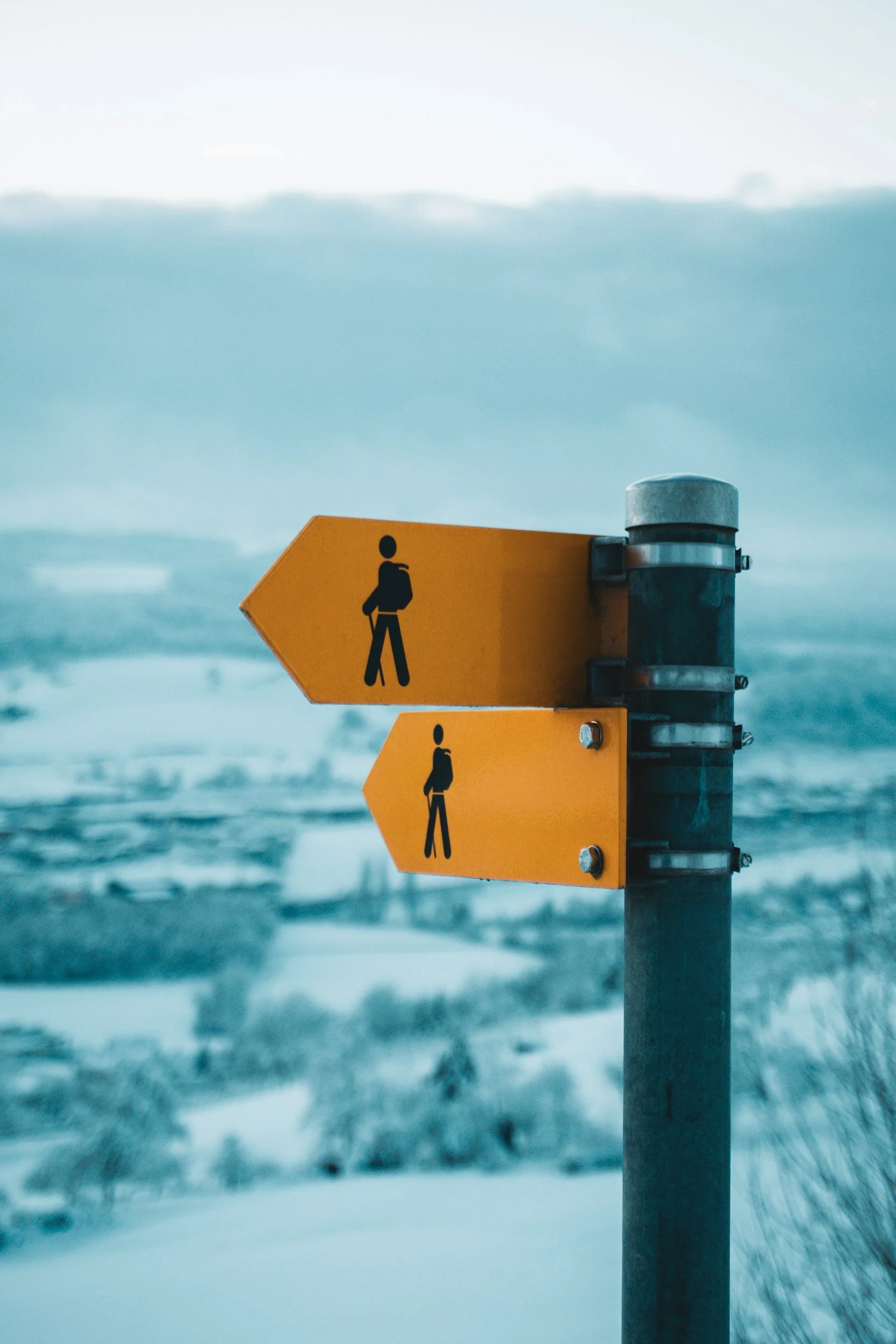 Two yellow hiking trail signs with black walking icons, one pointing left and the other straight, against a snowy landscape with hills and trees in the background.