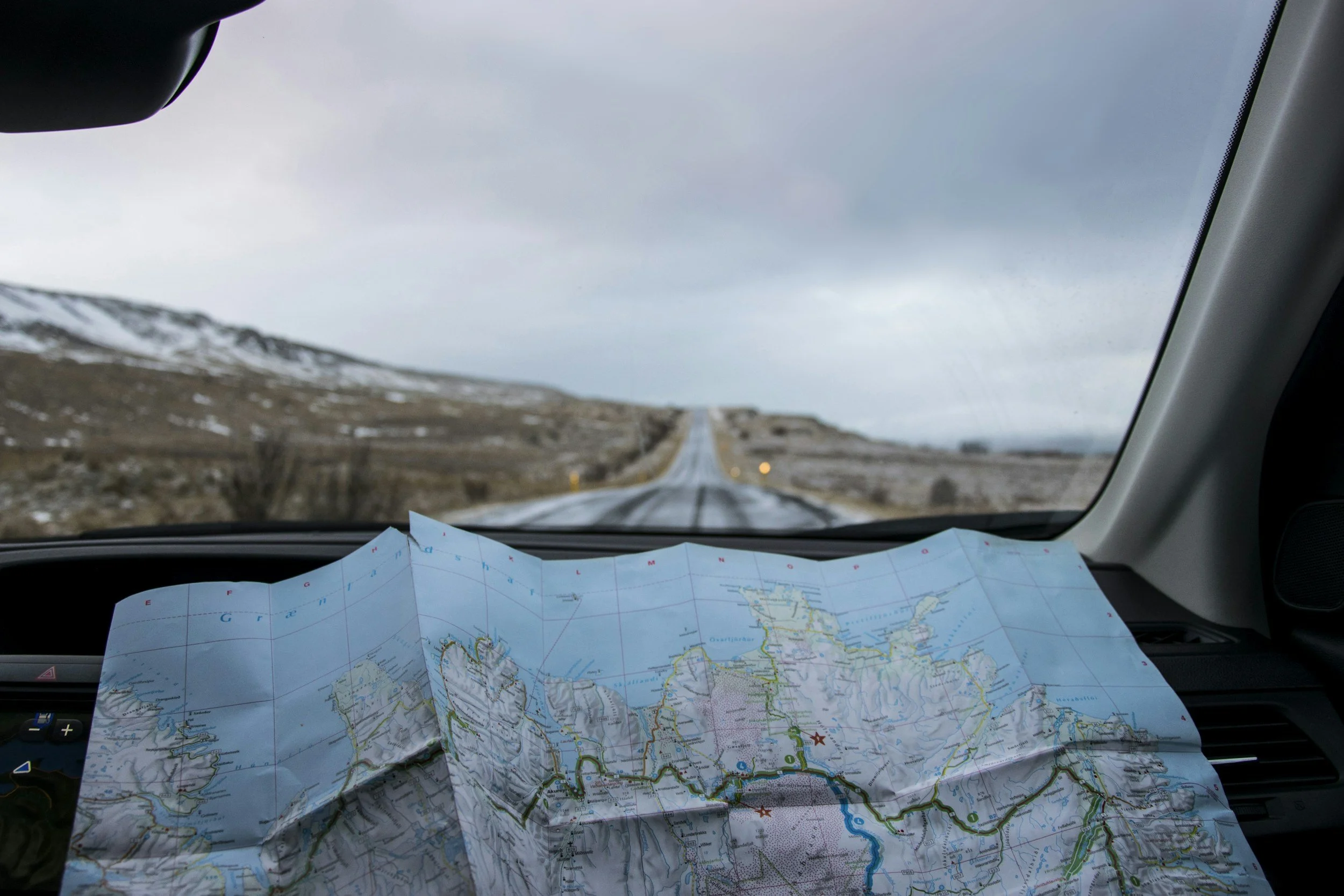 A map spread out on the dashboard of a car showing a road trip route with a rural landscape and cloudy sky through the windshield.