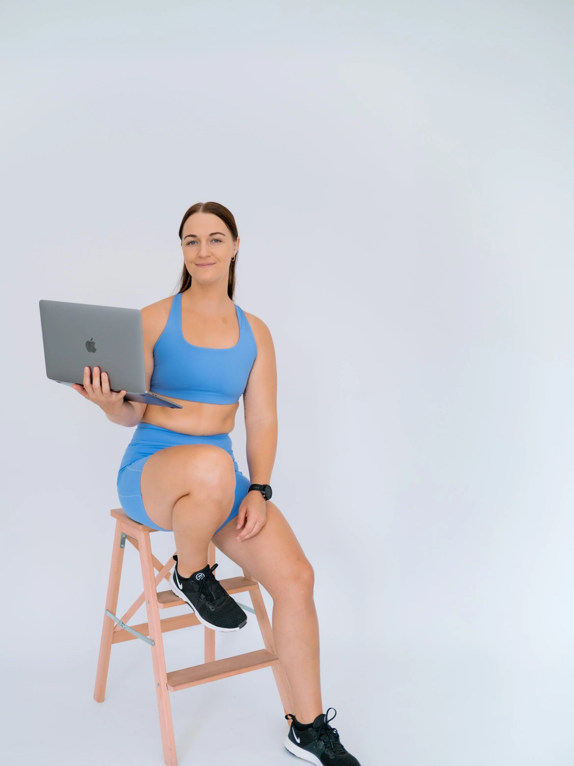 A woman in workout attire sitting on a wooden step stool holding a laptop in her left hand, against a plain white background.
