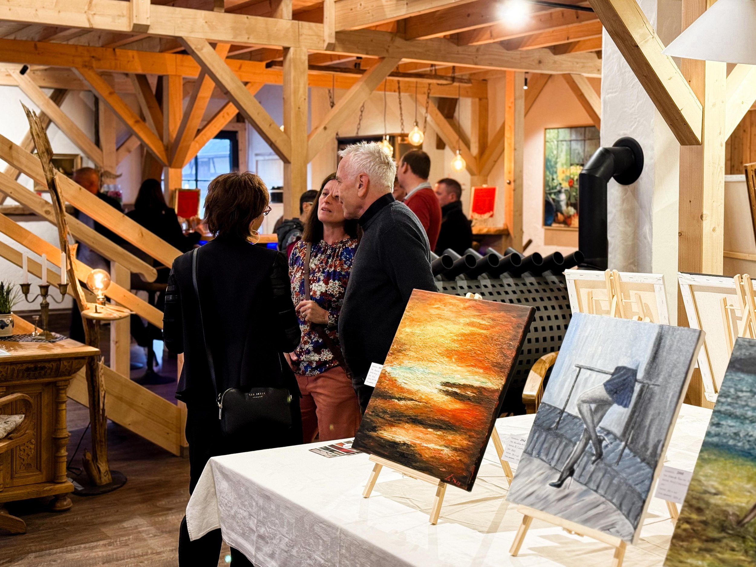 Menschen sprechen in einer Kunstgalerie, im Vordergrund sind Gemälde auf Tischen, im Hintergrund ist die Galerie mit Holzrahmen und Beleuchtung sichtbar.