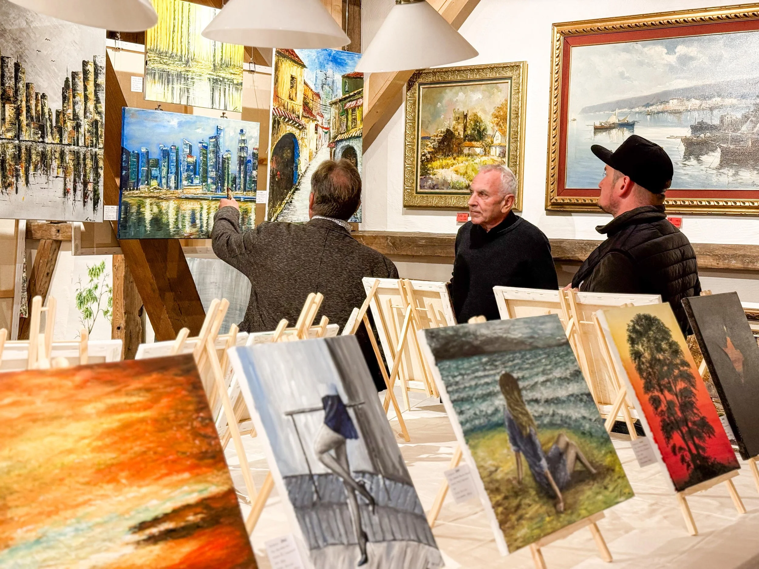 Drei Männer in einem Kunstausstellungsraum, die sich Gemälde ansehen, mit mehreren aufgestellten und an der Wand hängenden Gemälden, darunter Stadtansichten, Landschaften und abstrakte Kunst.