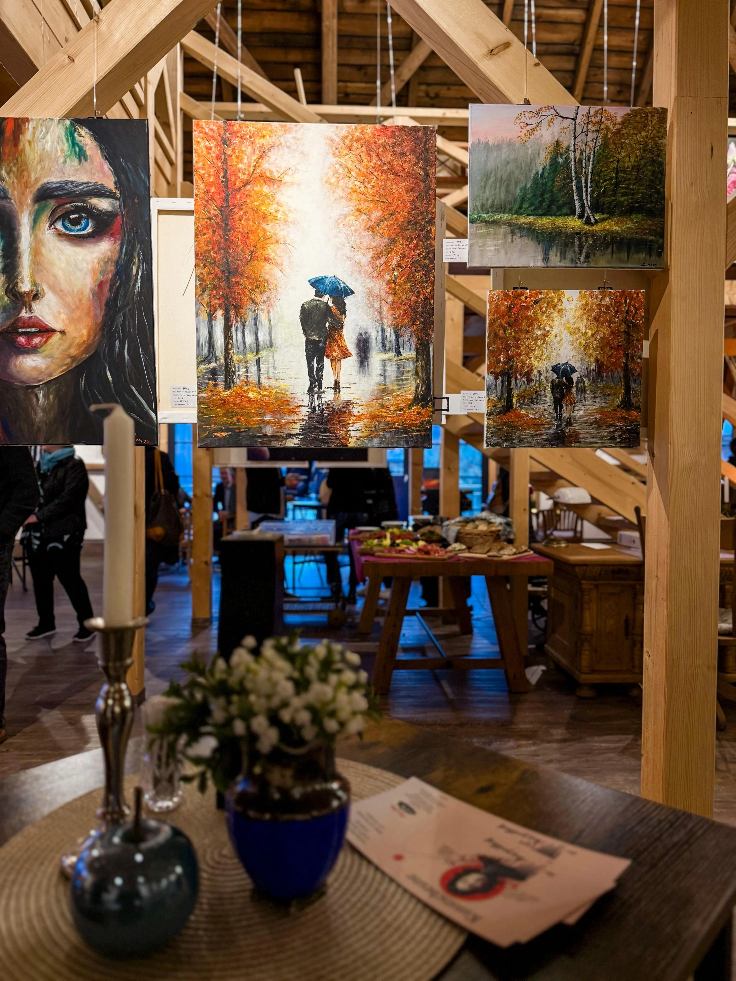 Verschiedene Gemälde in einem Kunstgalerie- oder Ausstellungsraum, darunter eine Portraitmalerei, eine Szene mit Paar unter Regenschirm, eine Landschaft mit Wasser und Bäumen, und eine weitere Szene mit Paar unter Regenschirm, alles in lebendigen Far