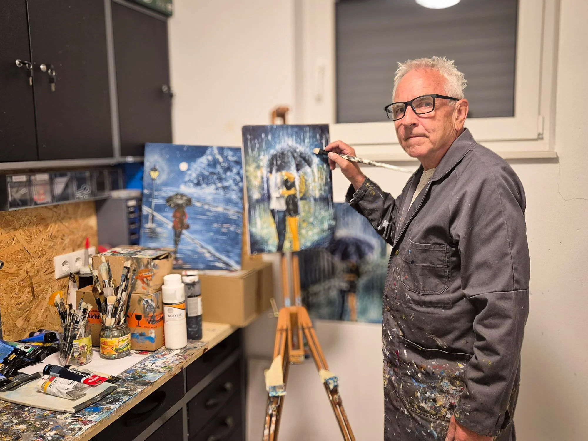 Ein älterer Mann in einer Kunstwerkstatt, der an einem Gemälde arbeitet. Es sind Malutensilien auf einem Tisch und zwei fertige oder halbfertige Leinwände zu sehen.