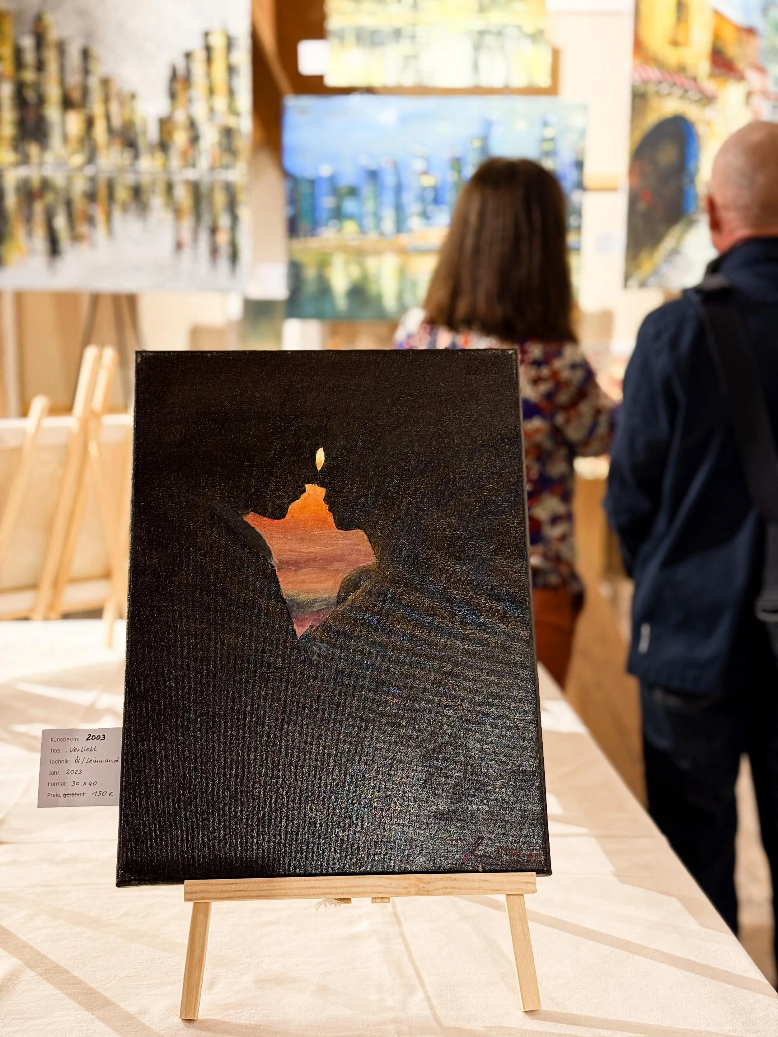 Kunstwerk auf einem kleinen Ständer, das eine Kerze in einer dunklen Szenerie zeigt, mit einem kleinen Licht am oberen Rand. Im Hintergrund sind zwei Personen, die Kunstwerke betrachten, und mehrere Gemälde sind an der Wand zu sehen.