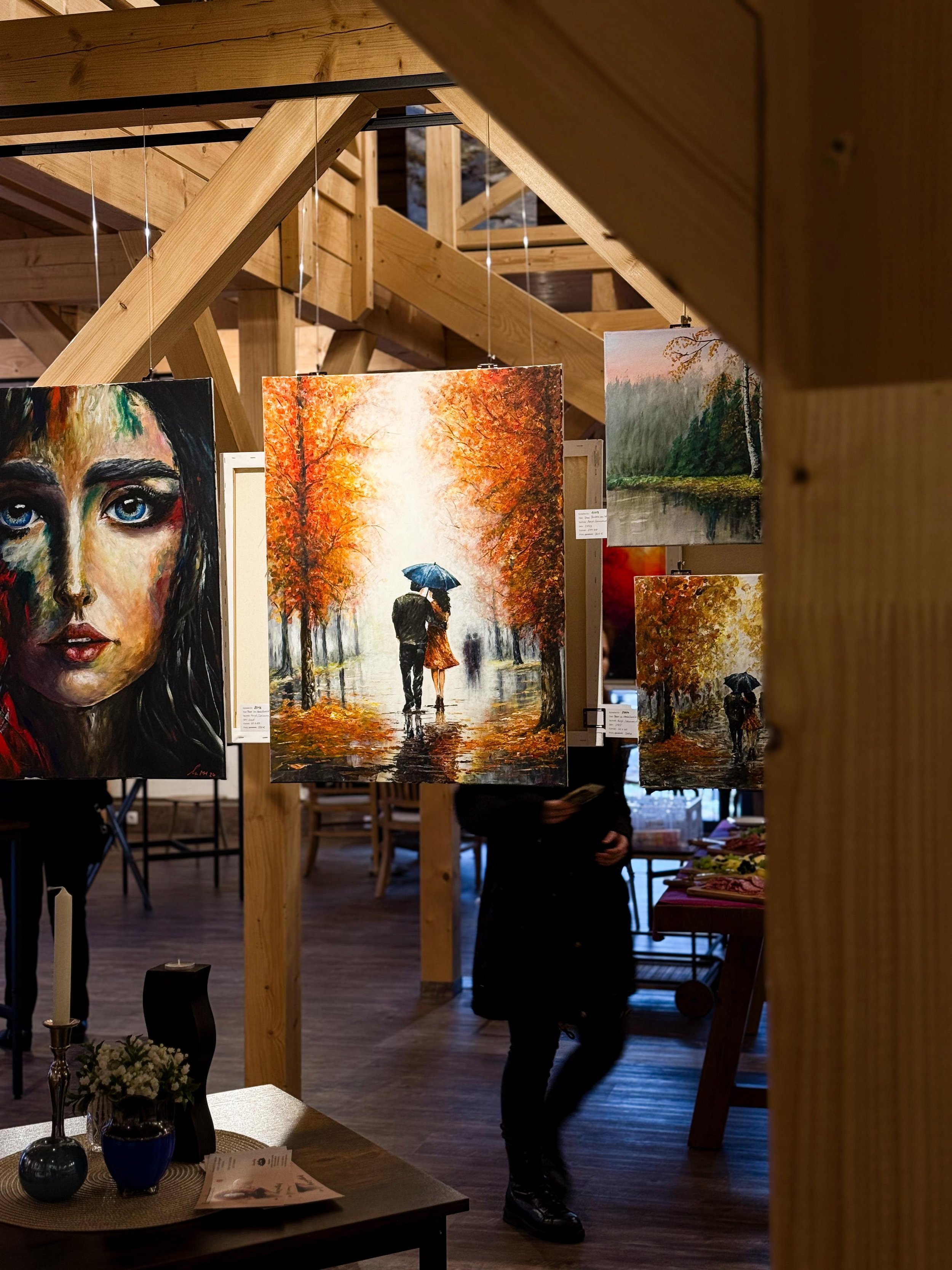 Gemälde in einer Kunstgalerie, darunter ein Porträt einer Frau mit leuchtenden blauen Augen und ein sommerliches Landschaftsbild mit einem Paar unter einem Regenschirm auf einem regenverzierten Weg, umgeben von bunten Herbstbäumen.