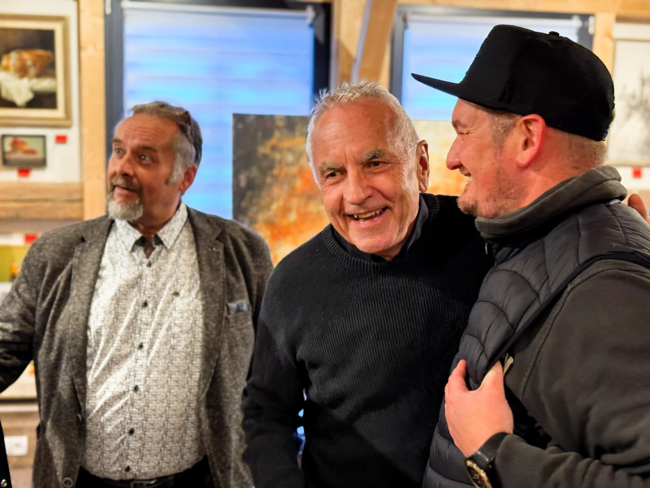 Drei Männer stehen in einem Raum mit Kunstwerken an der Wand. Zwei Männer lächeln und unterhalten sich, während der dritte Mann im Hintergrund ebenfalls lächelt.