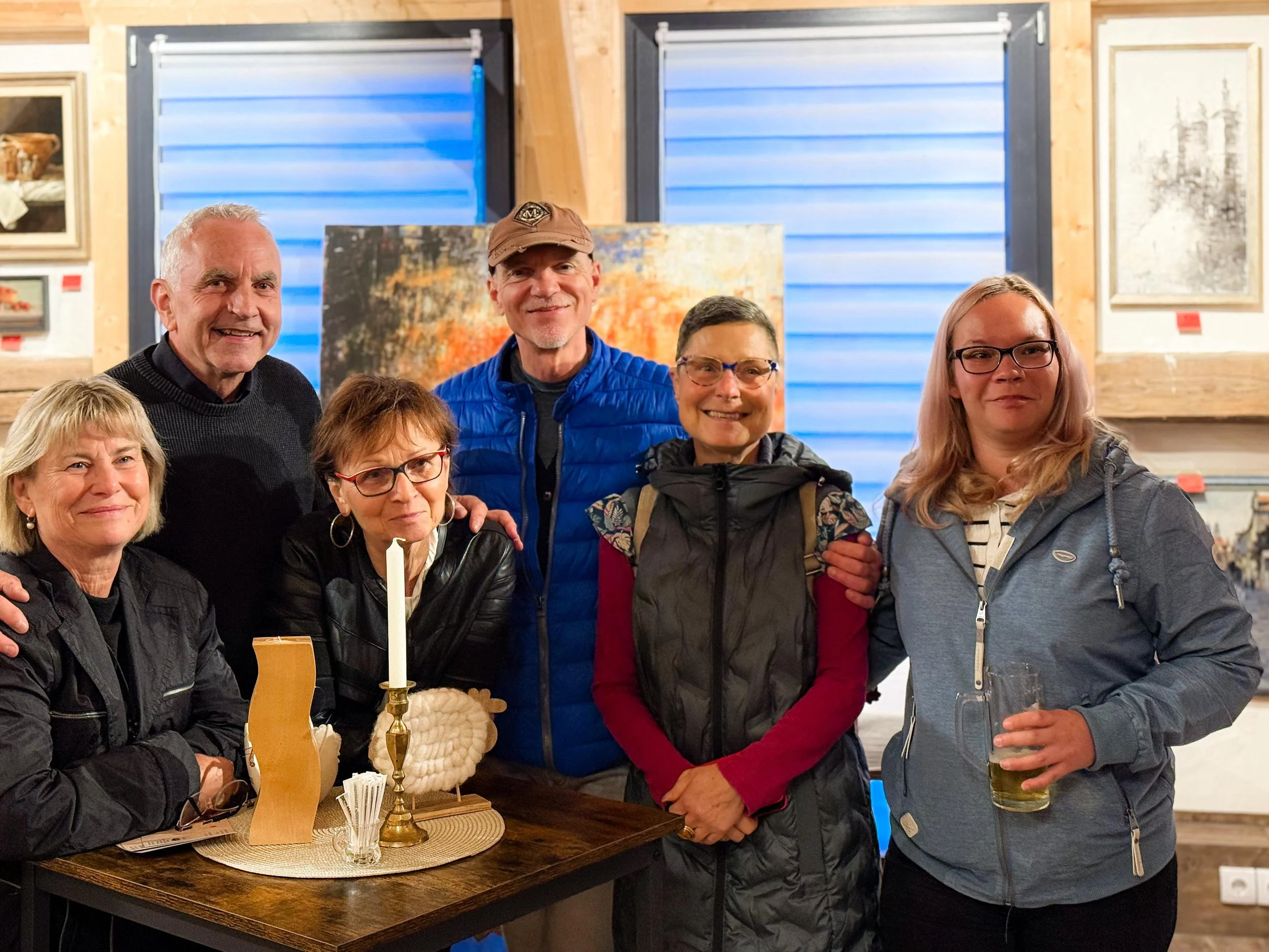 Eine Gruppe von sechs Menschen, fünf Frauen und ein Mann, posieren in einer Kunstgalerie. Sie sind lächelnd und tragen Sommer- oder Übergangskleidung. Im Vordergrund steht ein Tisch mit einer Kerze, einem Kerzenhalter und einer dekorativen Vase.