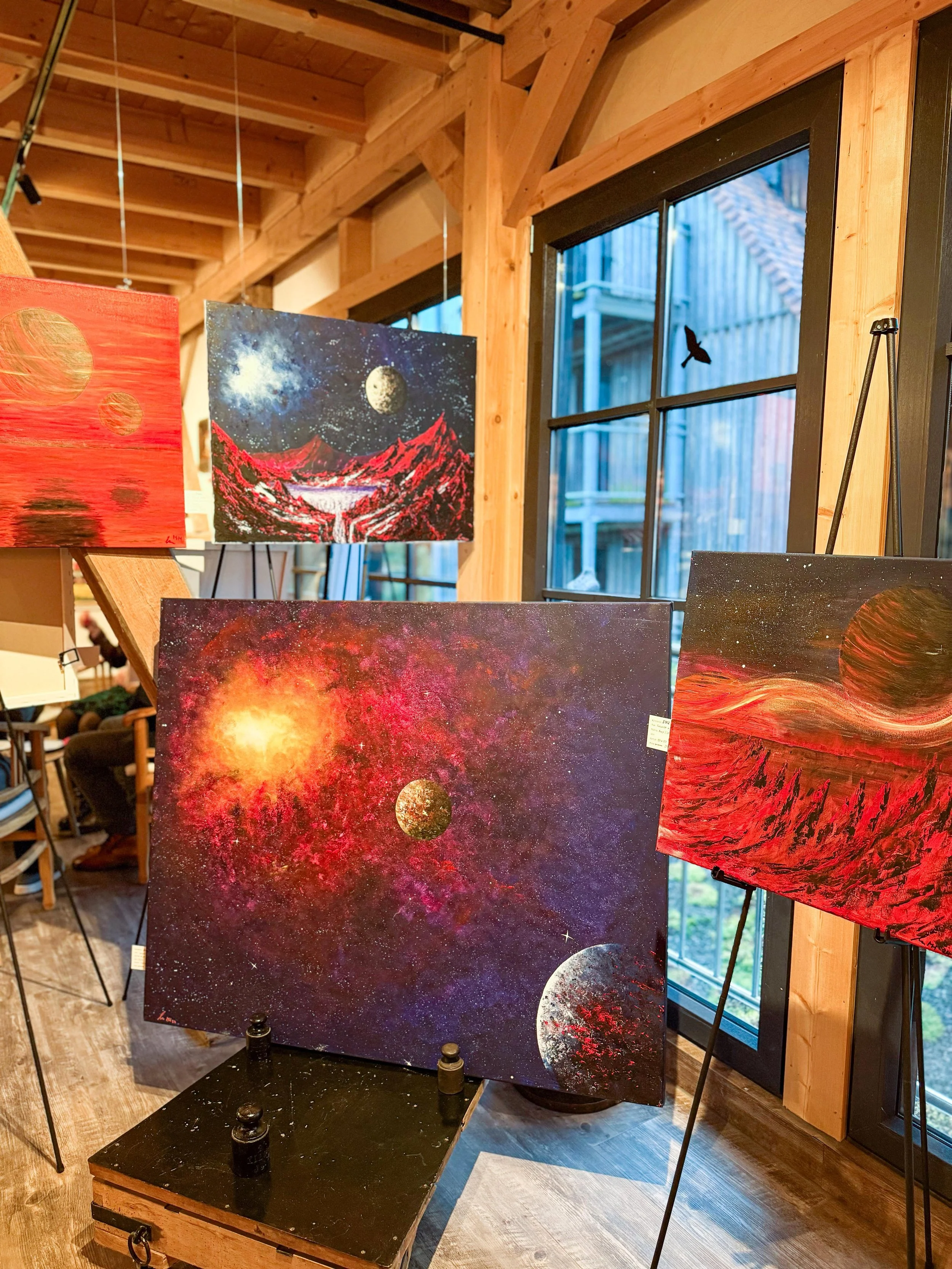 Ausstellung mit gemalten Bildern von Raum- und Planetenkonstellationen in einem Holzgebäude.