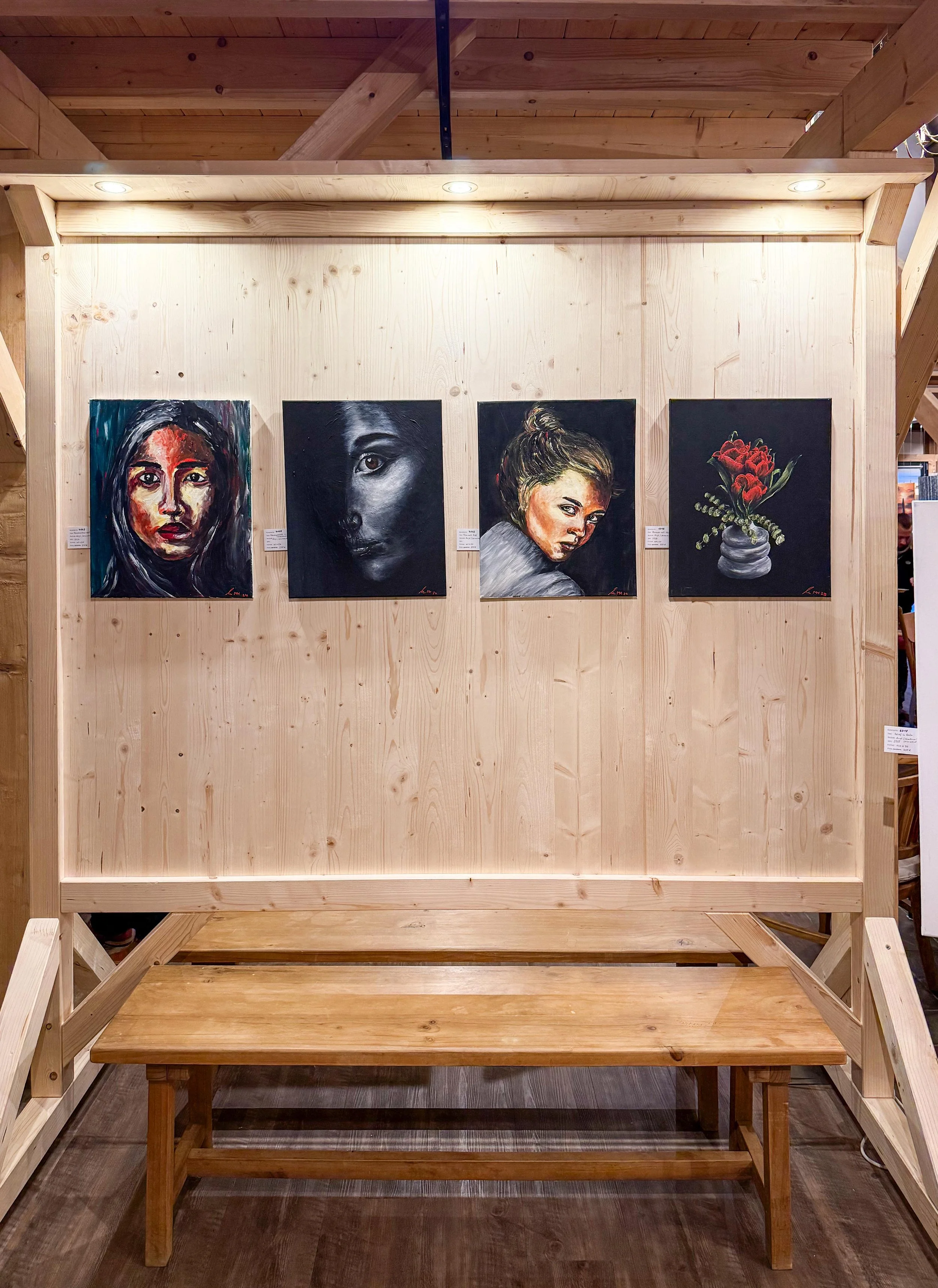 Ein Holzrahmen mit vier Gemälden von Frauen und einer Vase mit roten Rosen, beleuchtet von Deckenstrahlern, in einem Museum oder einer Galerie.