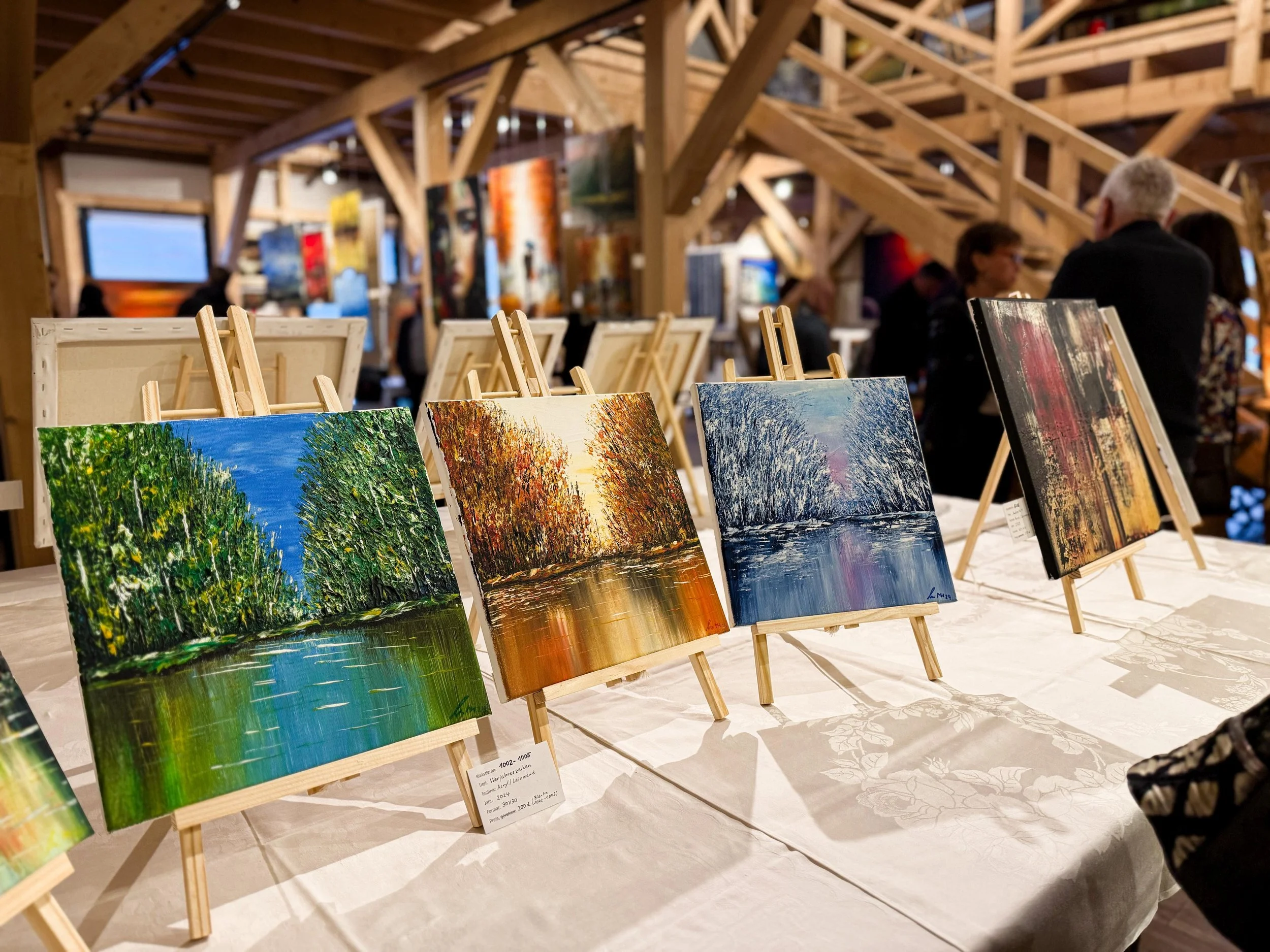 Vier kleine Leinwände mit impressionistischen Landschaftsbildern auf Tischen in einem Kunstmarkt, im Hintergrund Menschen und weitere Kunstwerke in einem Holzgebäude.