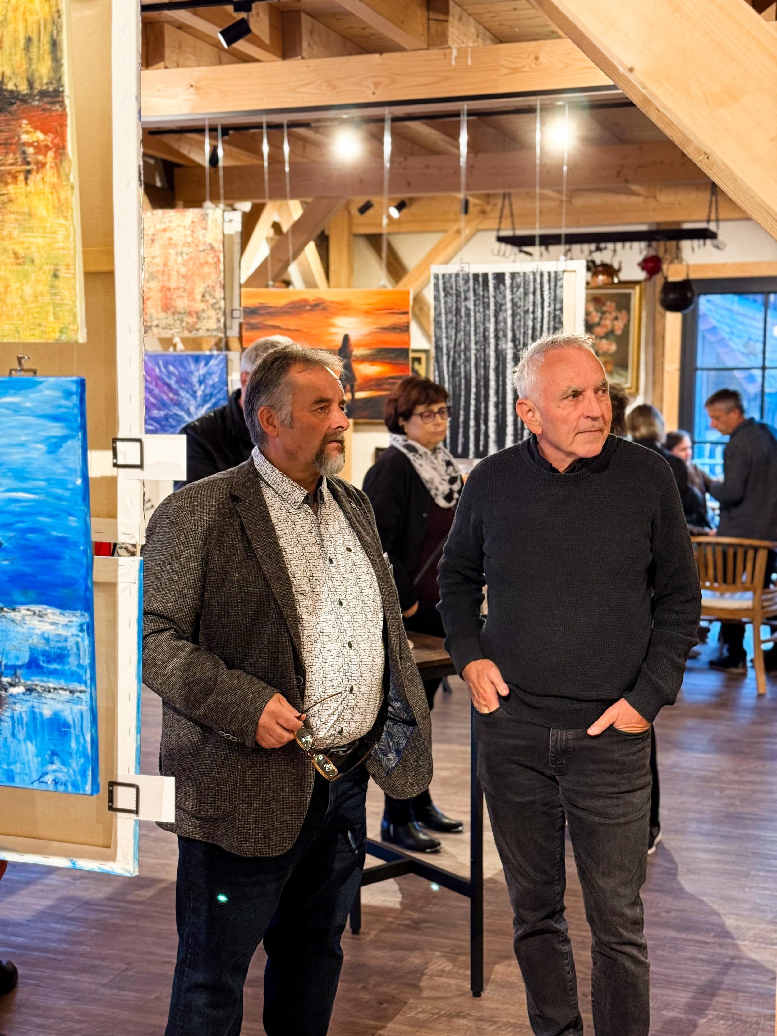 Zwei Männer stehen in einem Kunstgalerie, umgeben von Gemälden, einige Männer und Frauen sind im Hintergrund, die meisten blicken auf die Kunstwerke.