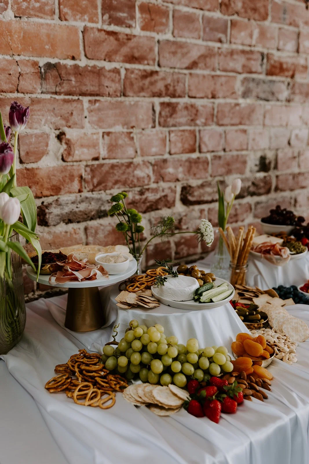 A cheese and fruit platter with grapes, strawberries, and dried apricots, along with crackers, bread, and assorted deli meats on a white tablecloth against a brick wall.