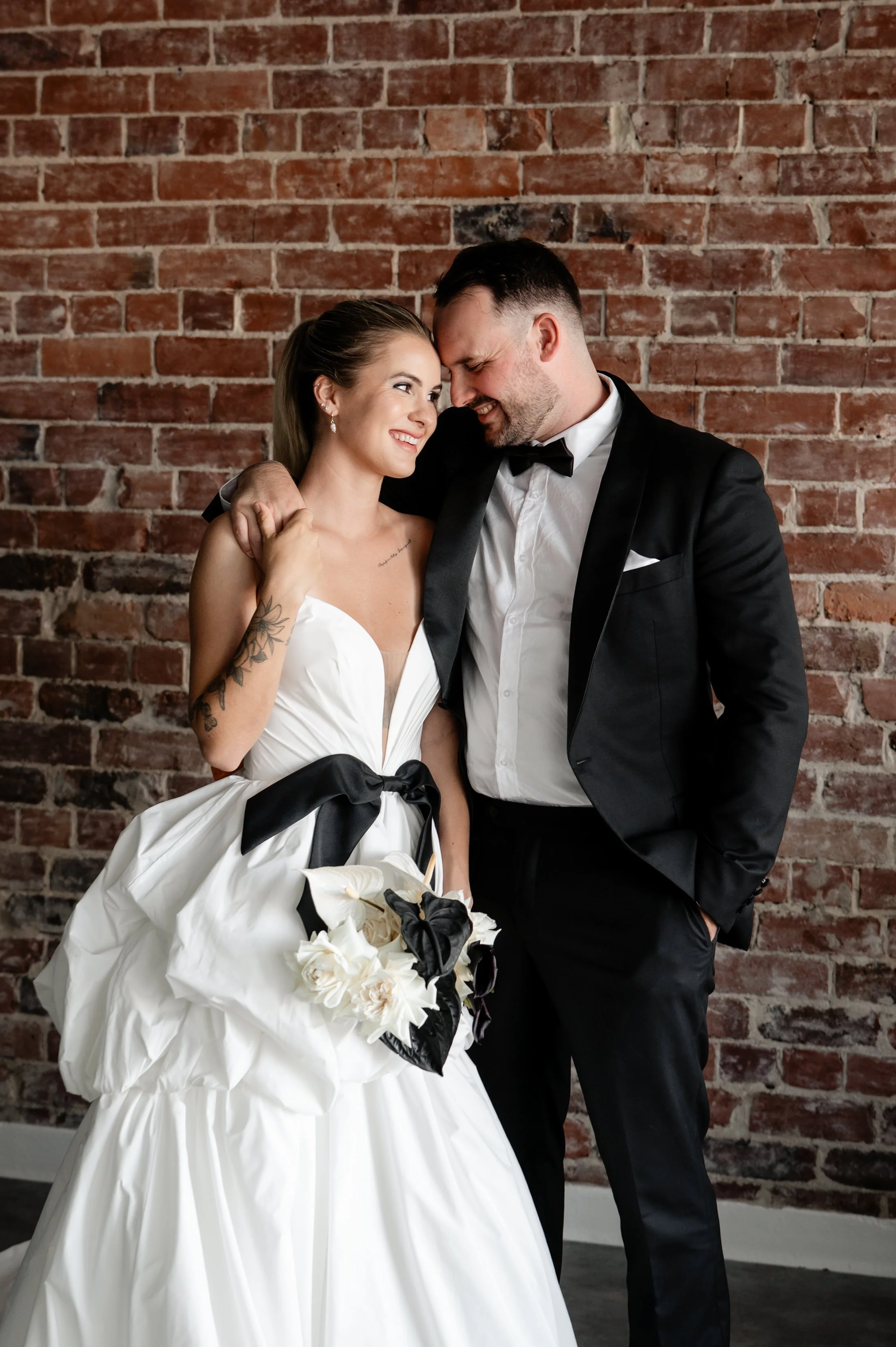 A bride and groom smiling affectionately, standing close together against a brick wall. The bride is in a white wedding dress with a black ribbon and holding a bouquet, and the groom is in a black tuxedo with a black bow tie.