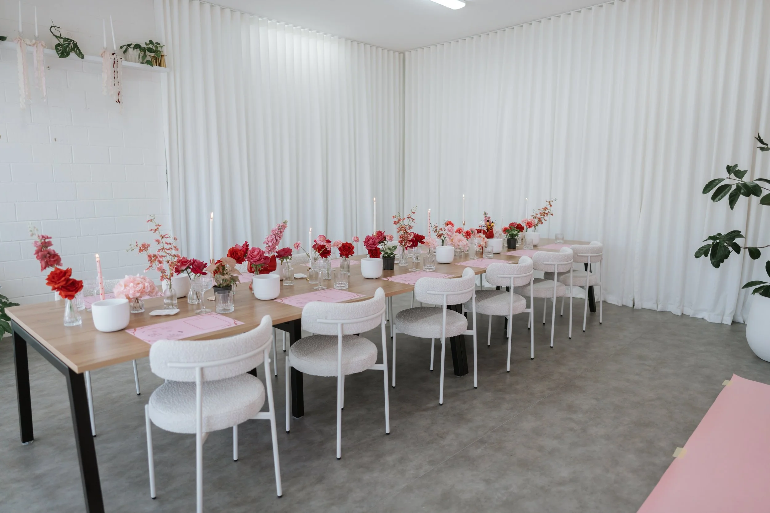 A long dining table decorated with pink and red flowers in glass vases, set for a celebration with pink placemats and tall pink candles in white holders. The room has white curtains, white chairs with rounded backs, potted plants, and a white brick wall.