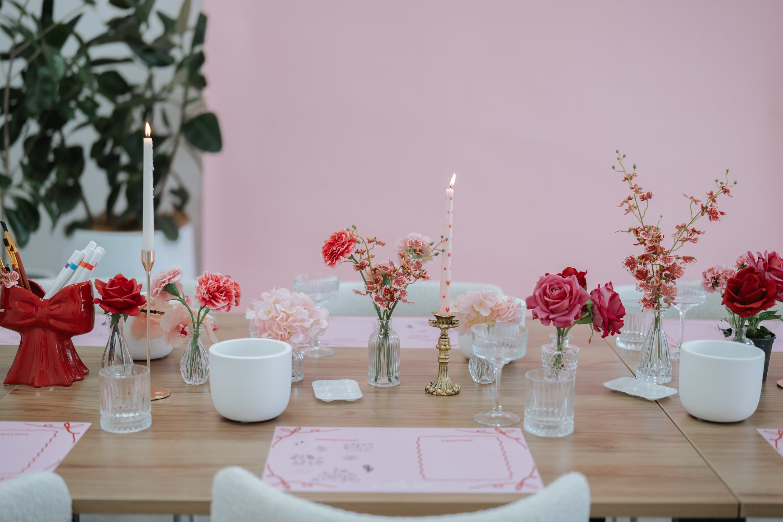 A decorated dining table with pink flowers, white and gold candles, and pink placemats, set against a pink wall with a green plant in the background.