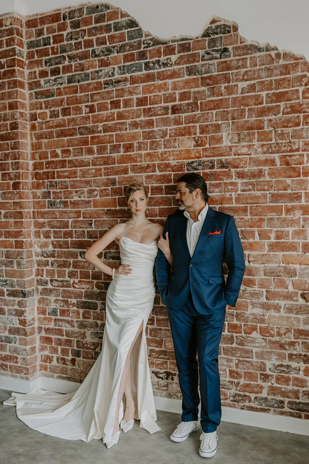 A woman in a strapless white wedding gown with a slit, and a man in a navy blue suit with white sneakers, standing against an exposed brick wall.