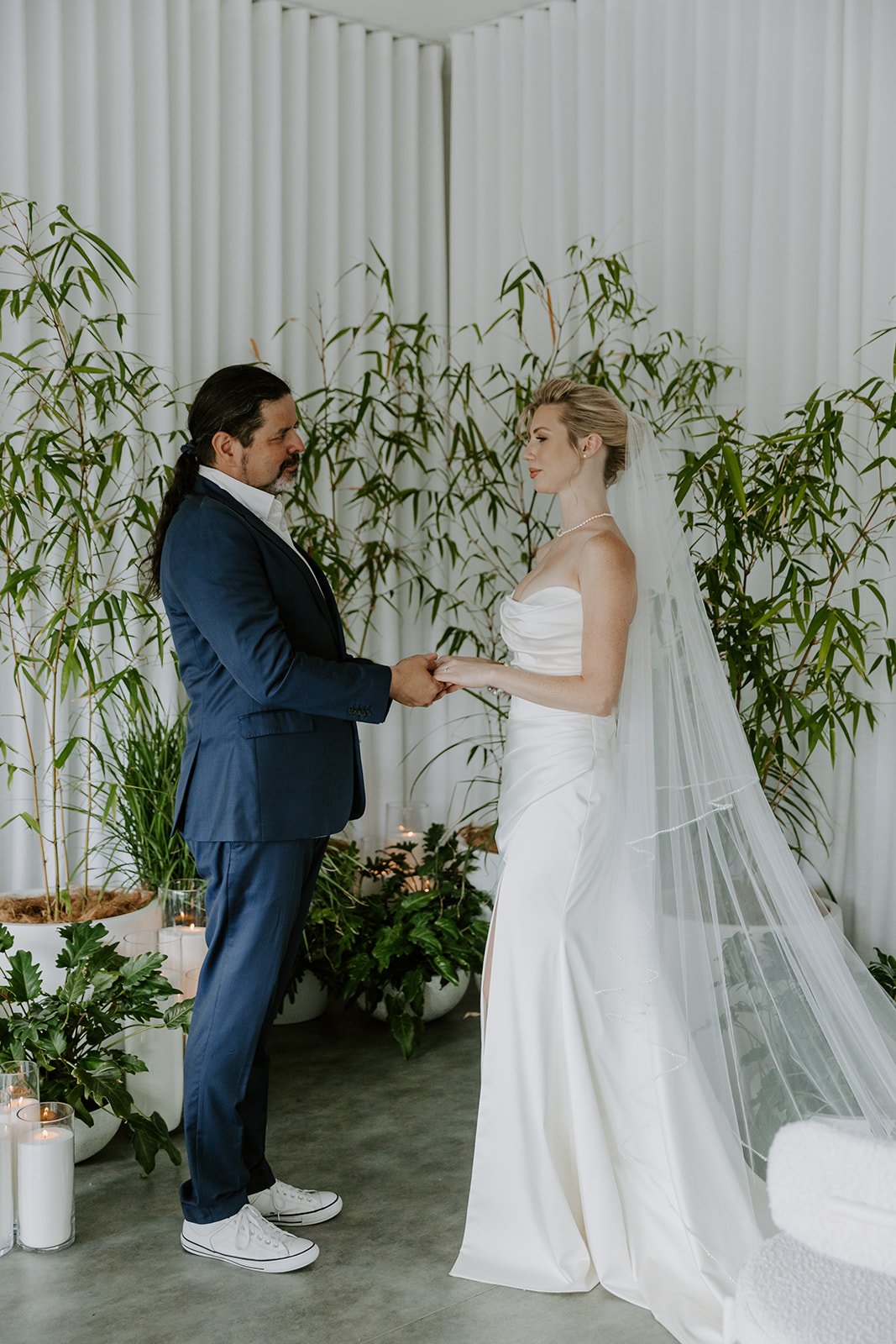 A couple exchanging vows during their wedding ceremony, with green plants and white curtains in the background.