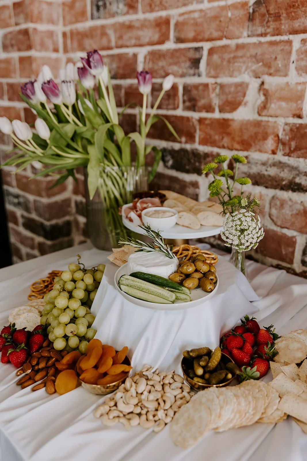 A table with cheese, grapes, strawberries, nuts, pickles, dried apricots, bread, and flowers against a brick wall.