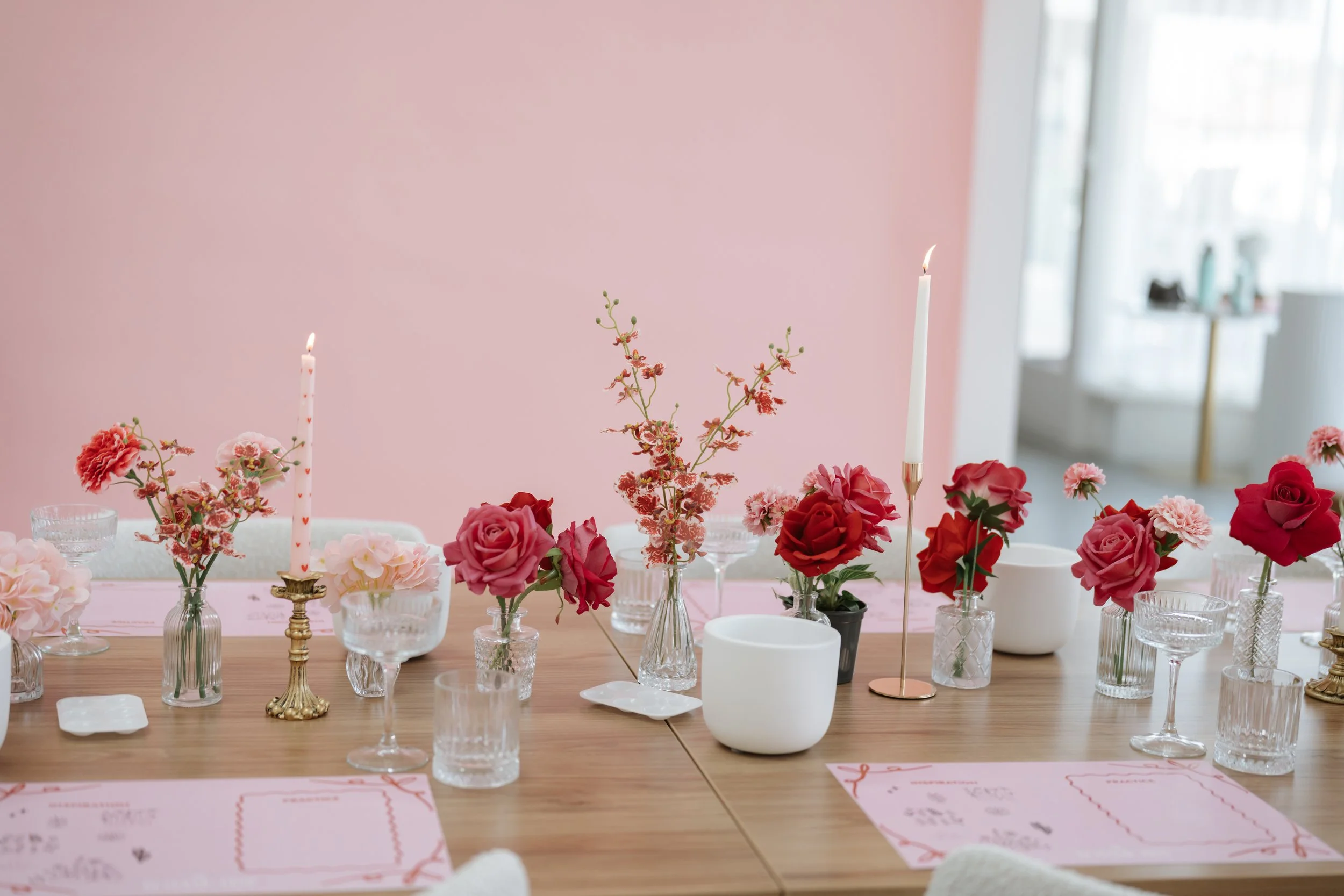 Table decorated with pink and red flowers in glass vases, white and gold candlesticks with lit candles, clear glassware, and pink paper placemats in a softly lit room with pink walls and large windows.