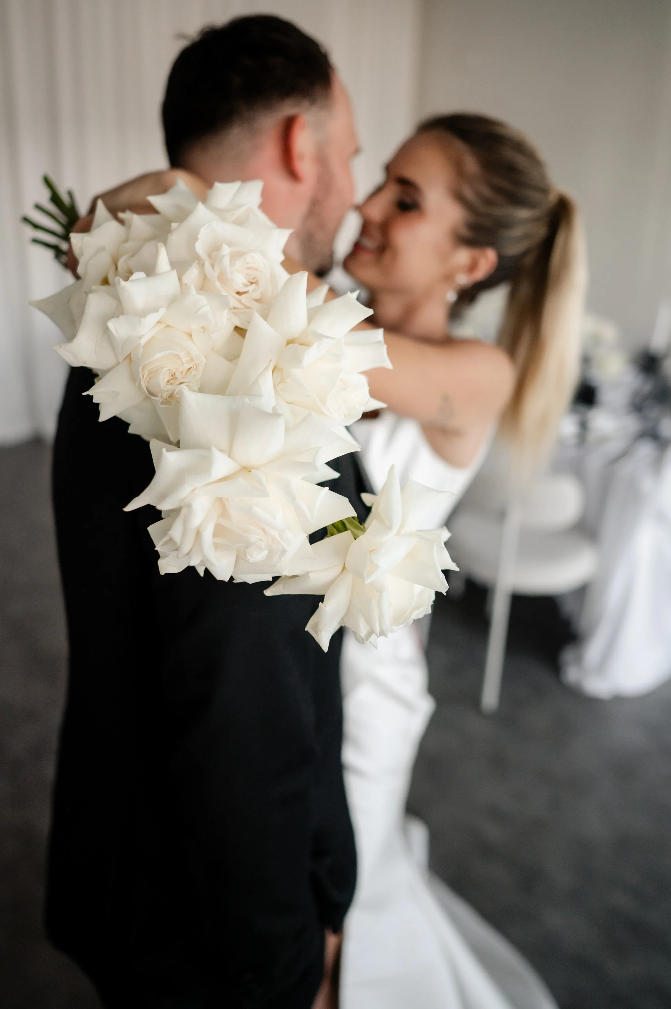 A bride and groom embrace, with the bride smiling at the groom. The bride is holding a bouquet of white roses. The scene is inside a decorated wedding venue.