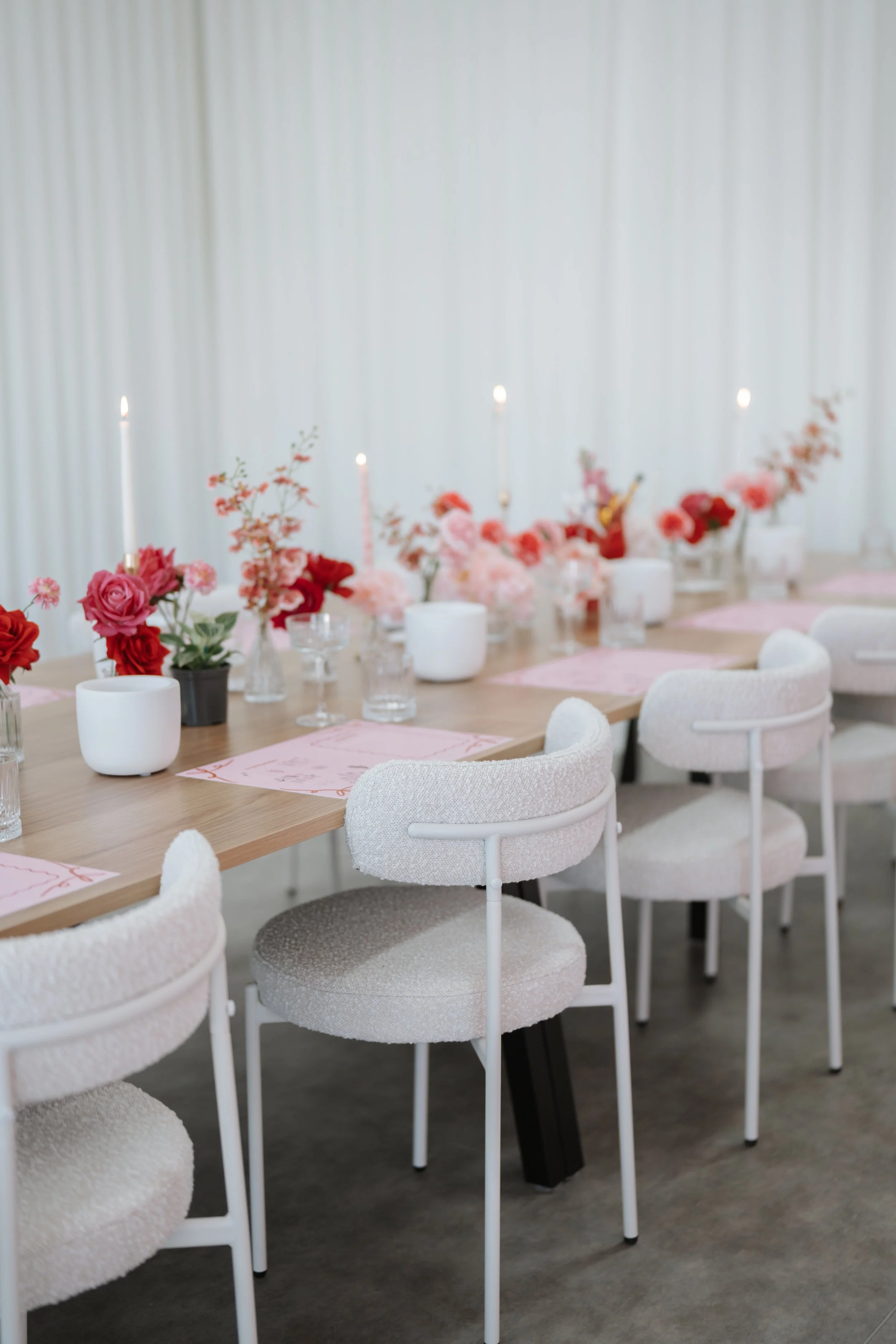 Decorated dining table with pink and red roses, candles, glassware, and pink place mats in a bright room with white curtains.