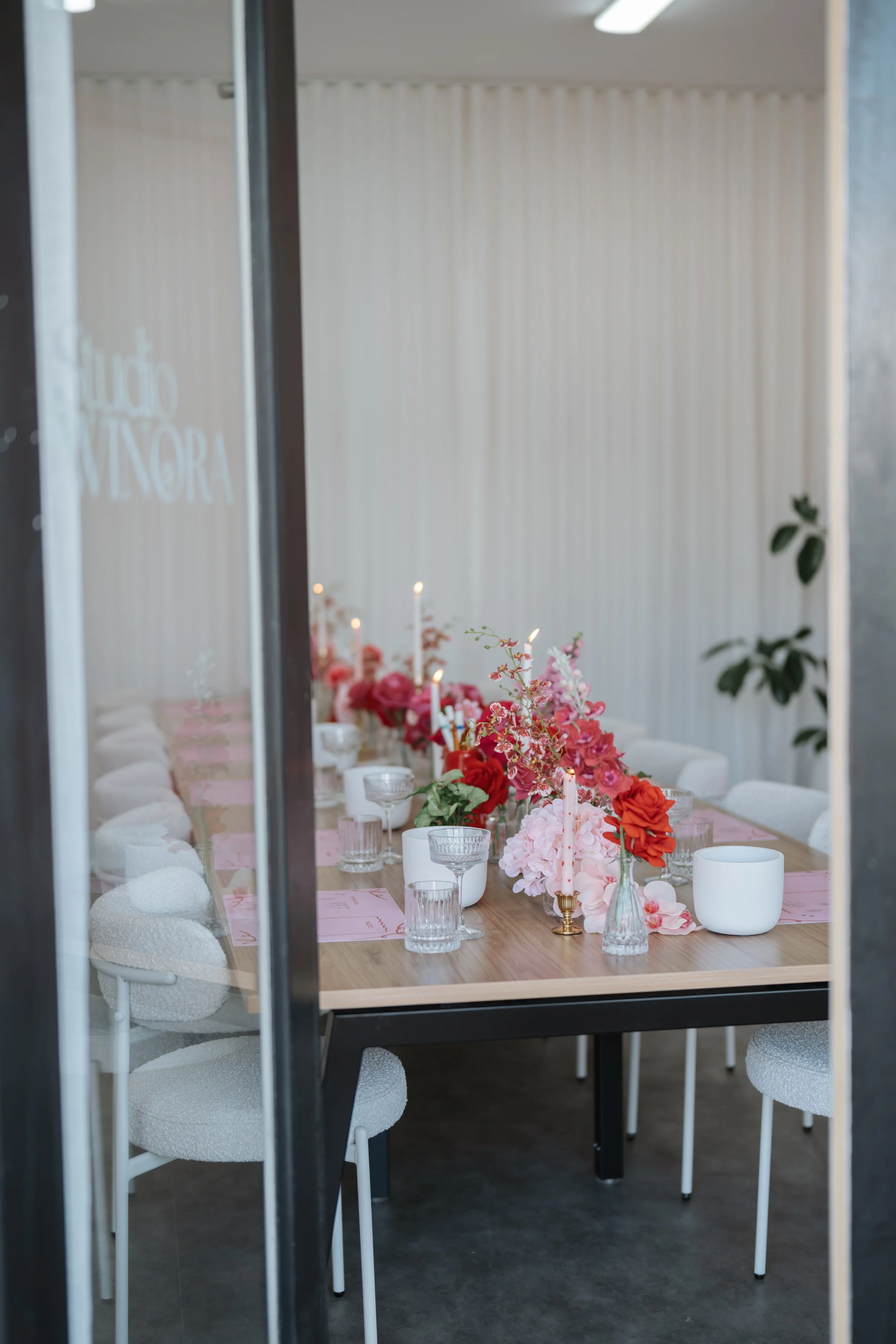 A long dining table decorated with pink and red flowers, candles, and glassware, viewed through a glass door with the words 'Studio Victoria' on it.