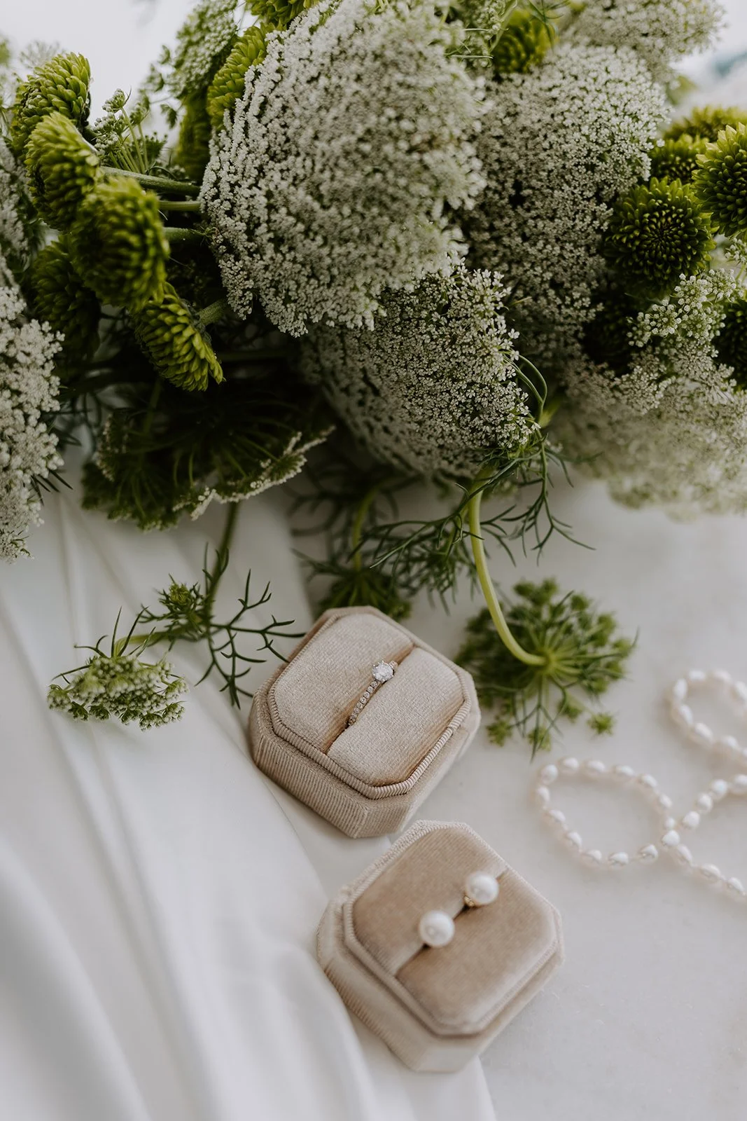 A bouquet of white and green flowers with a diamond engagement ring and pearl earrings displayed in beige velvet boxes on a white surface.