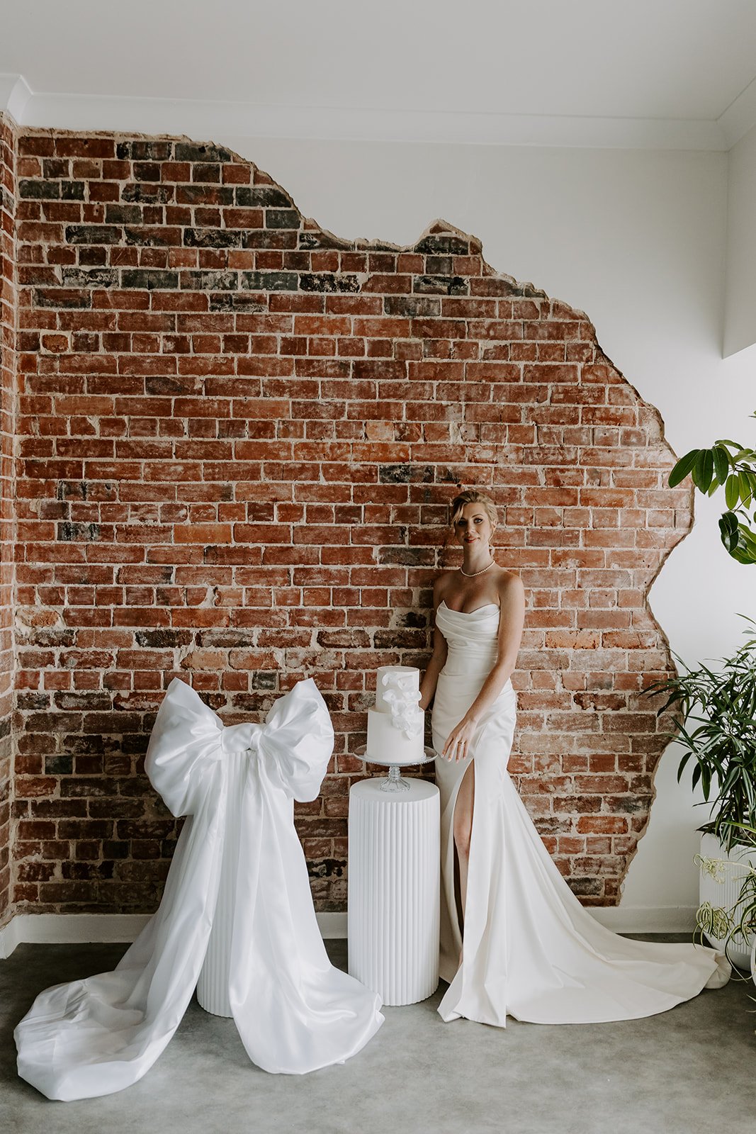 A woman in a white wedding gown standing next to a wedding cake on a pedestal, against an exposed brick wall. A large white satin bow is on the floor in front of her, and there are some green plants to the right.