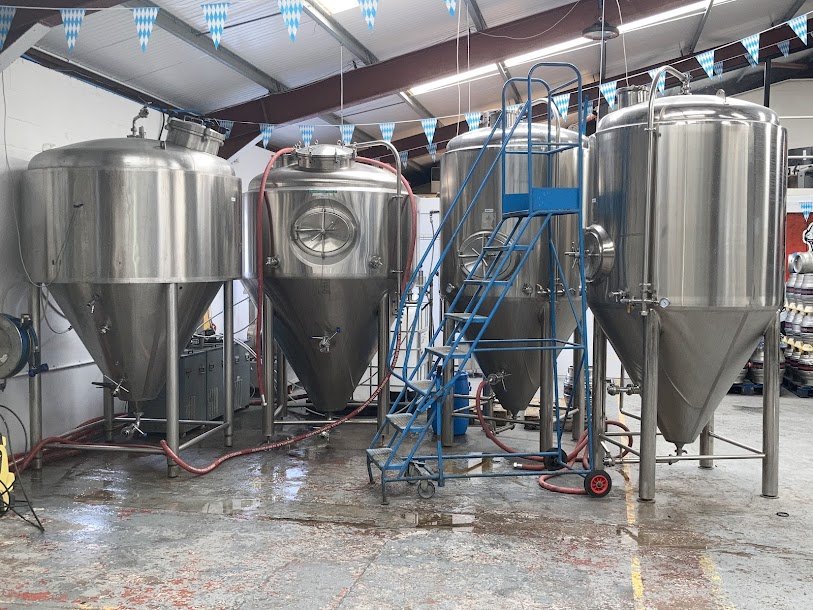 Industrial brewery equipment including stainless steel fermentation tanks and a blue staircase inside a brewery.